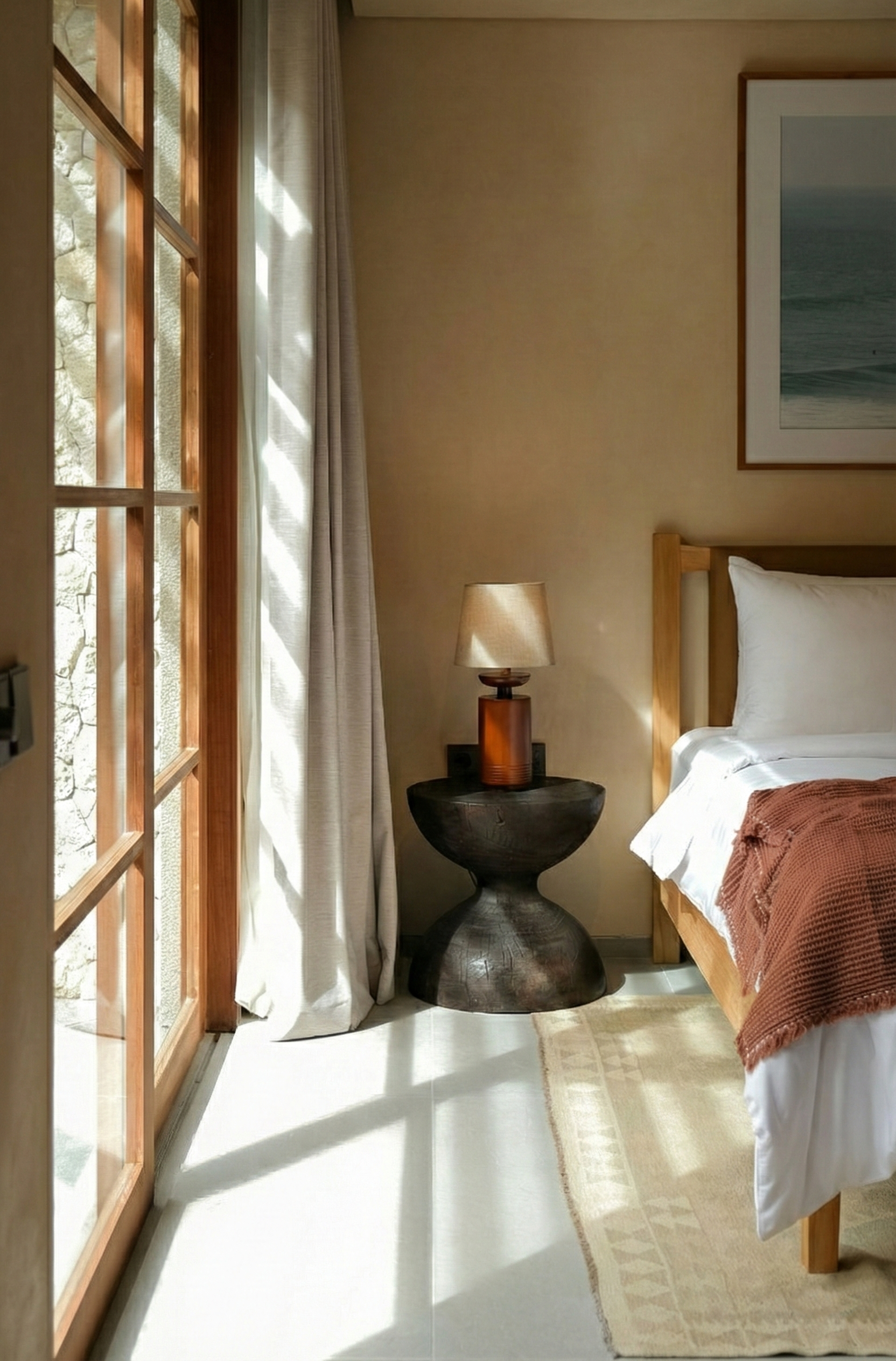 Sunlit bedroom corner with a wooden bed, bedspread, bedside table with a lamp, framed picture on the wall, and glass doors with curtains.