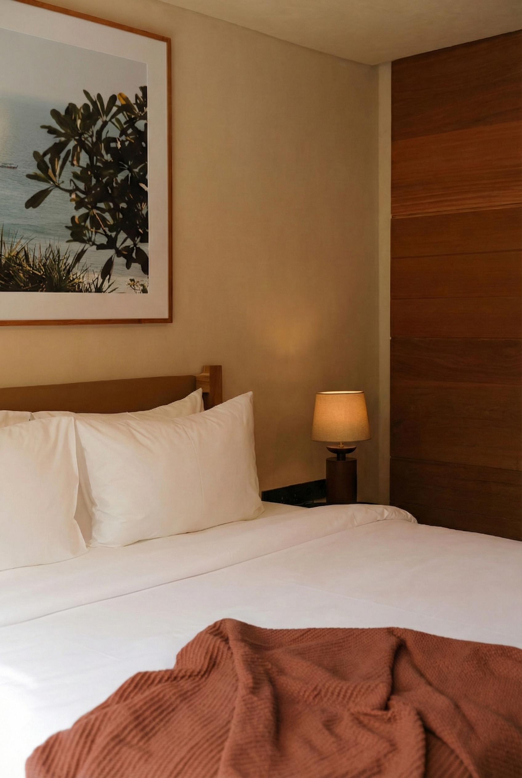 A hotel room with a bed, white pillows, a bedside lamp, a framed picture, and a brown blanket in the foreground.