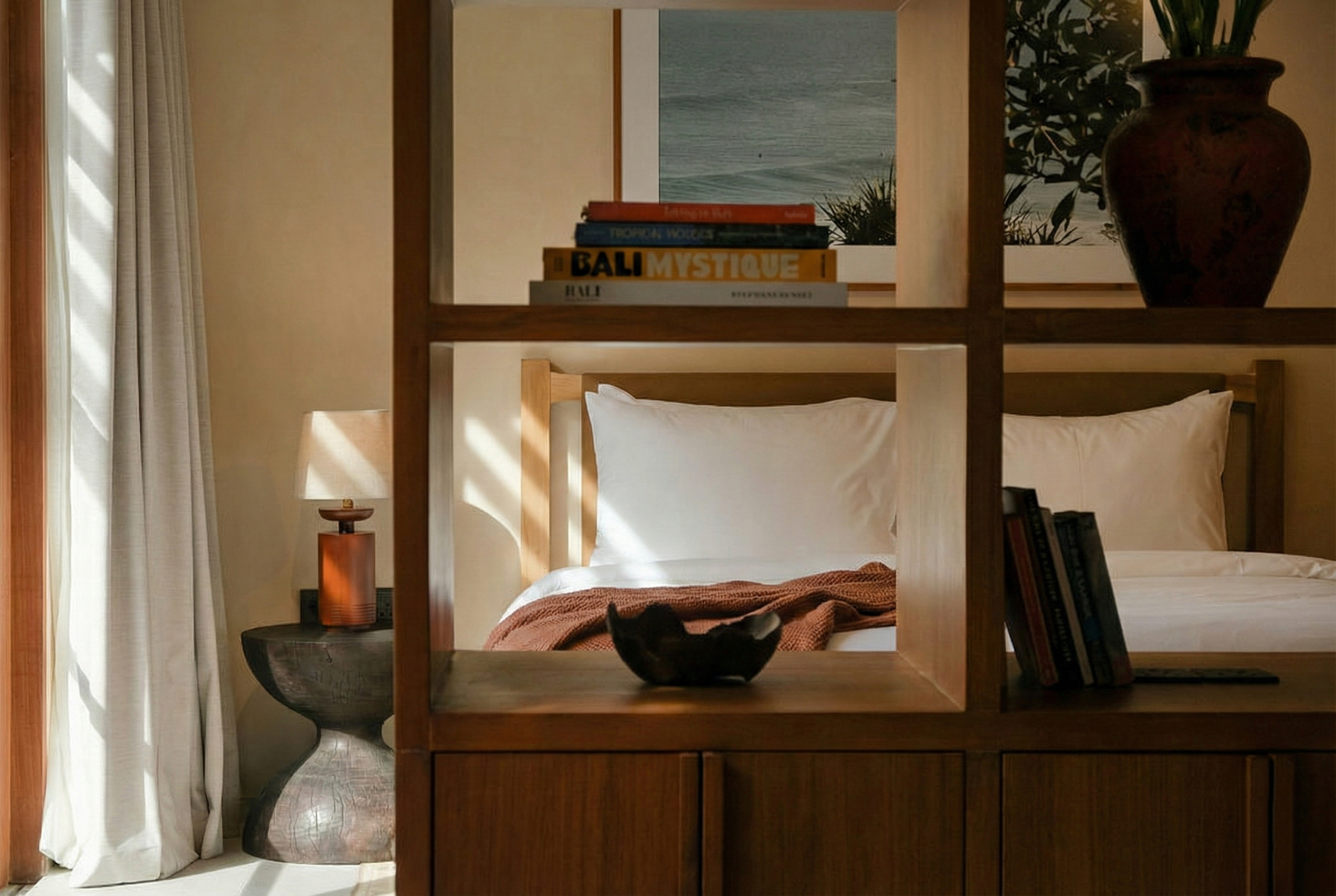 Interior of a cozy bedroom with a wooden shelf separating the beds. There is a small black cat in a bowl on the shelf, a table lamp, a stack of books, and framed artwork of the ocean on the wall.