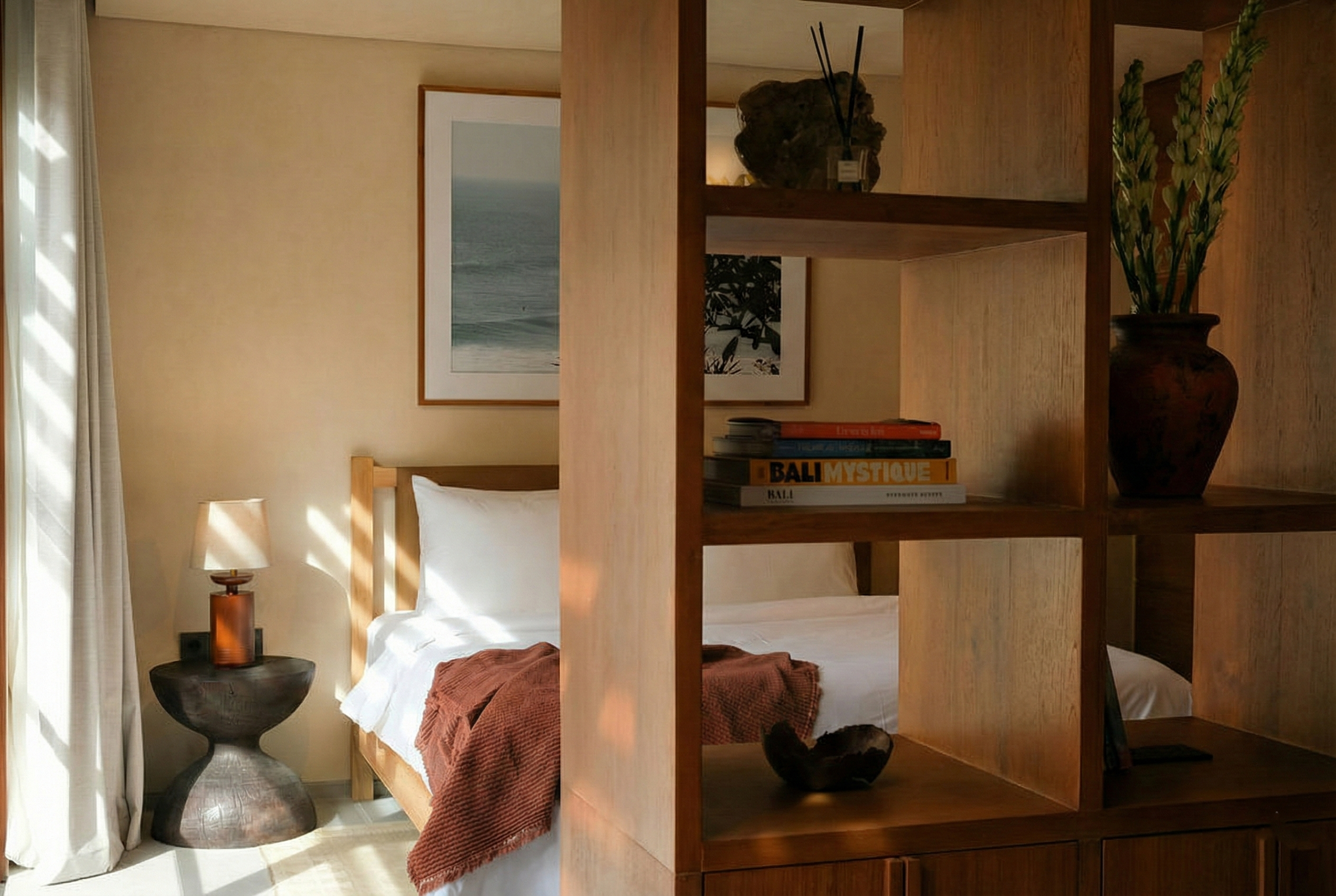 A bedroom with a bed and a wooden shelf divider. The bed has white bedding and a rust-colored throw blanket. Beside the bed, there is a dark wooden nightstand with a rounded base and a lamp with a brown shade. On the wall above the bed, there is fram