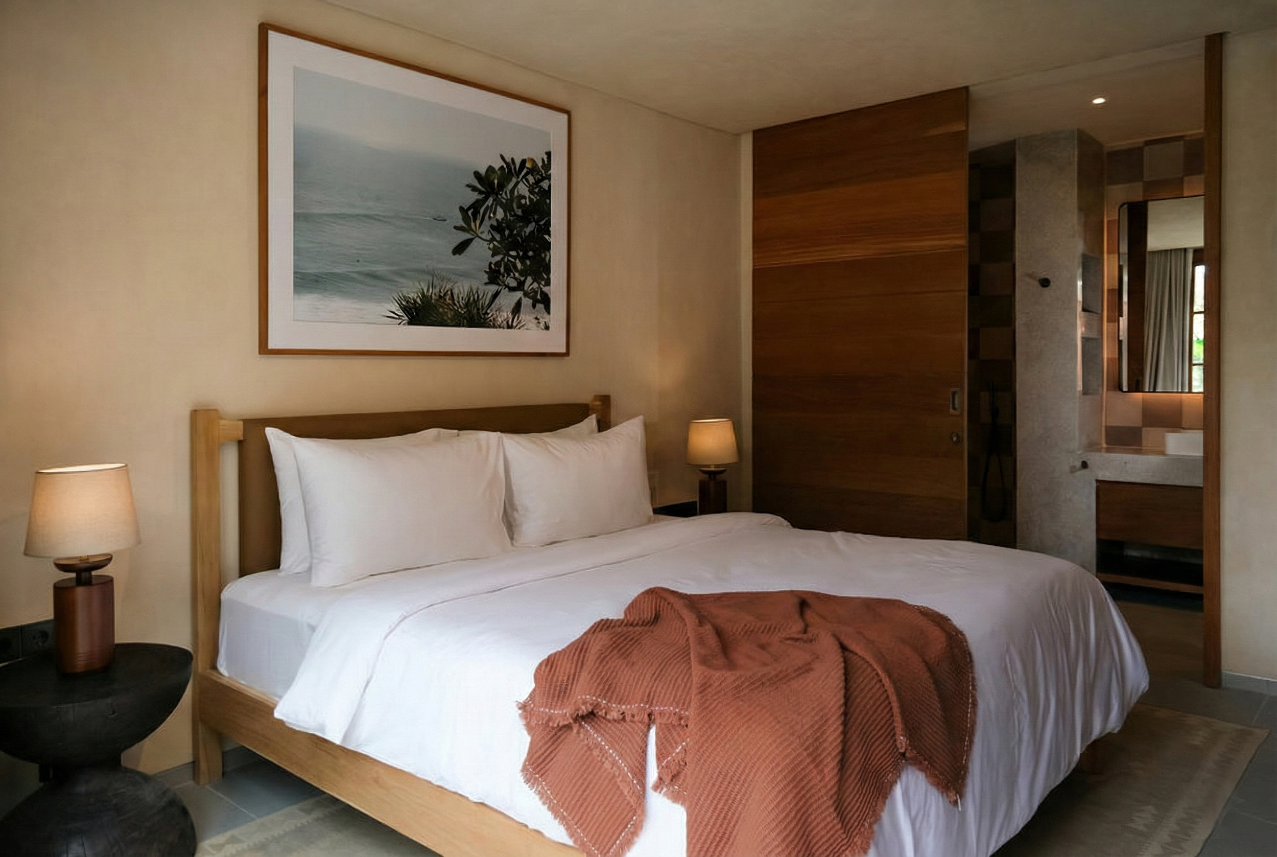 A hotel room with a bed, a painting of a seascape hanging above the headboard, two bedside lamps, and wooden partitions.