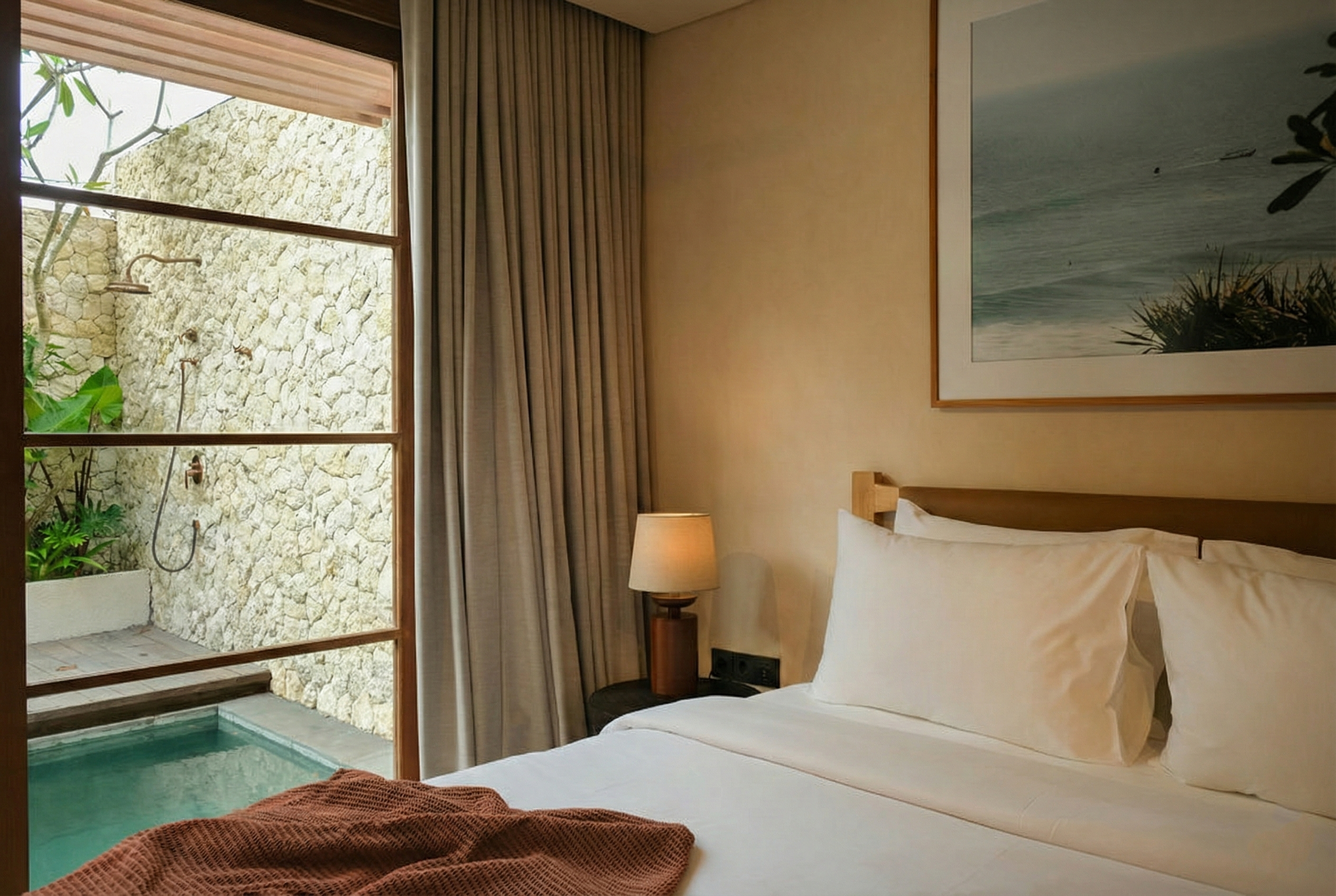 A hotel room with a large bed and white bedding, a wooden nightstand with a lamp, beige curtains by the window, and a framed beach scene painting on the wall. Outside the window, a stone wall, greenery, and outdoor shower with a stone floor can be se
