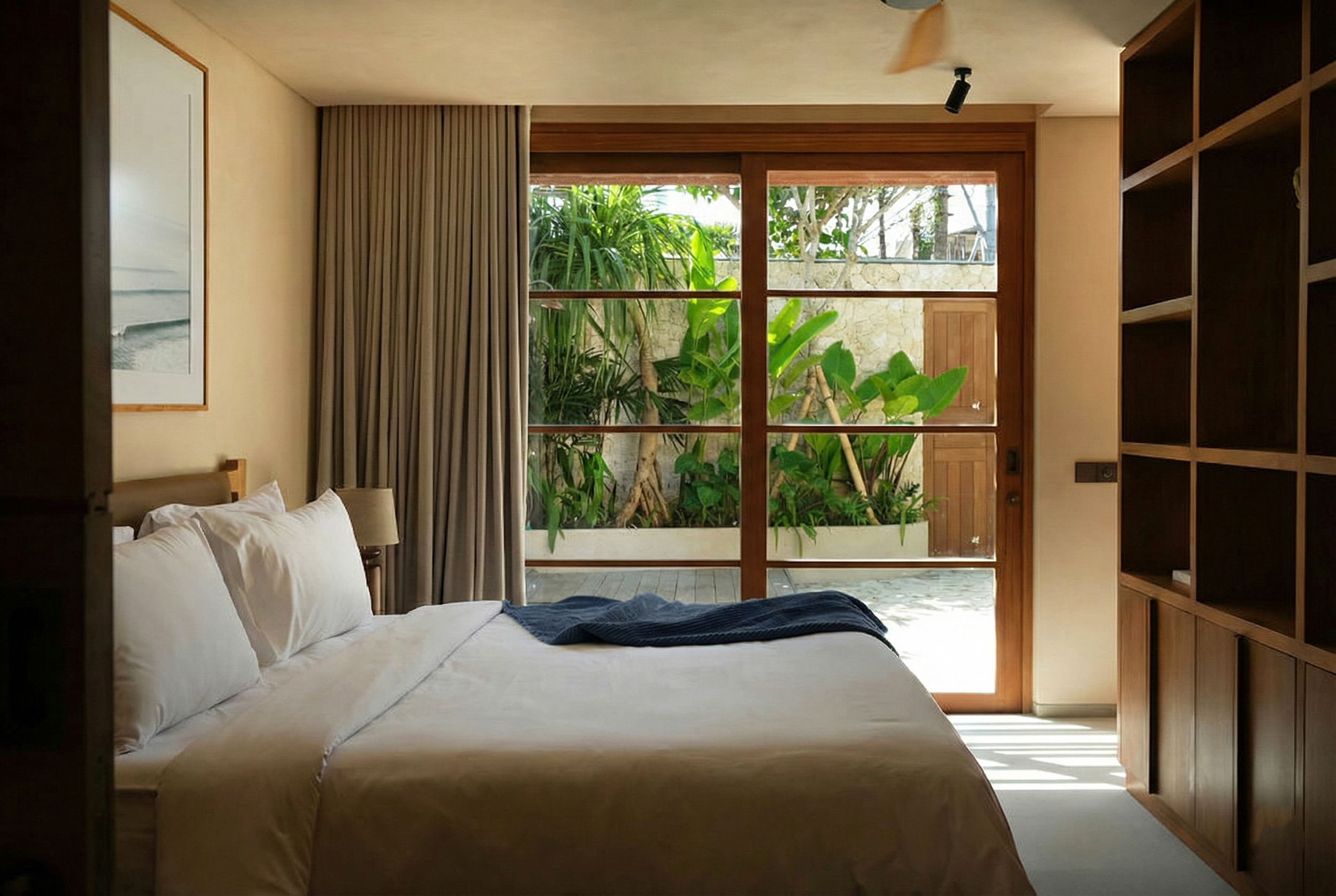 A bedroom with a large bed with white bedding and pillows, adjacent to a large window with wooden framing and view of green plants and sunlight outside.