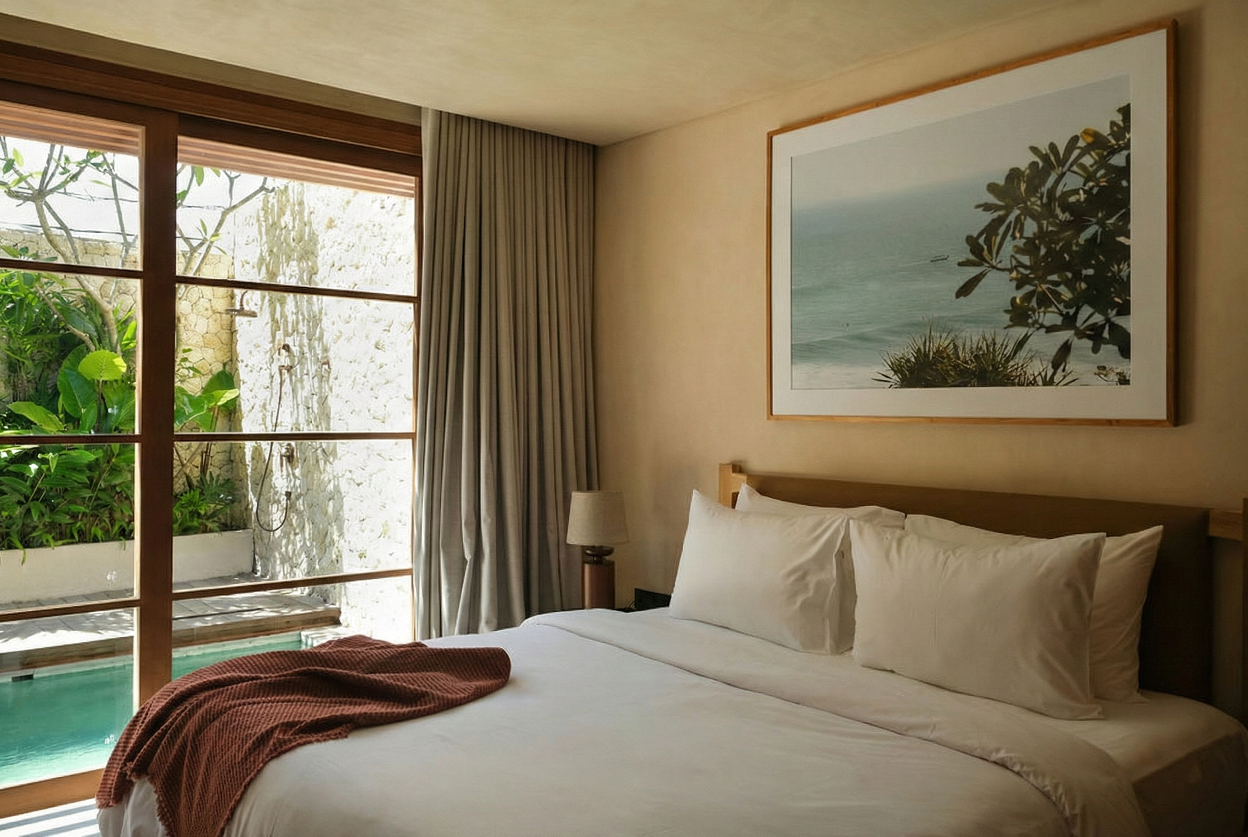 A bedroom with a large bed, white sheets, and pillows, featuring a picture of a seaside landscape on the wall. There is a window with wooden frames and a view of lush green plants outside, along with curtains hanging beside it.
