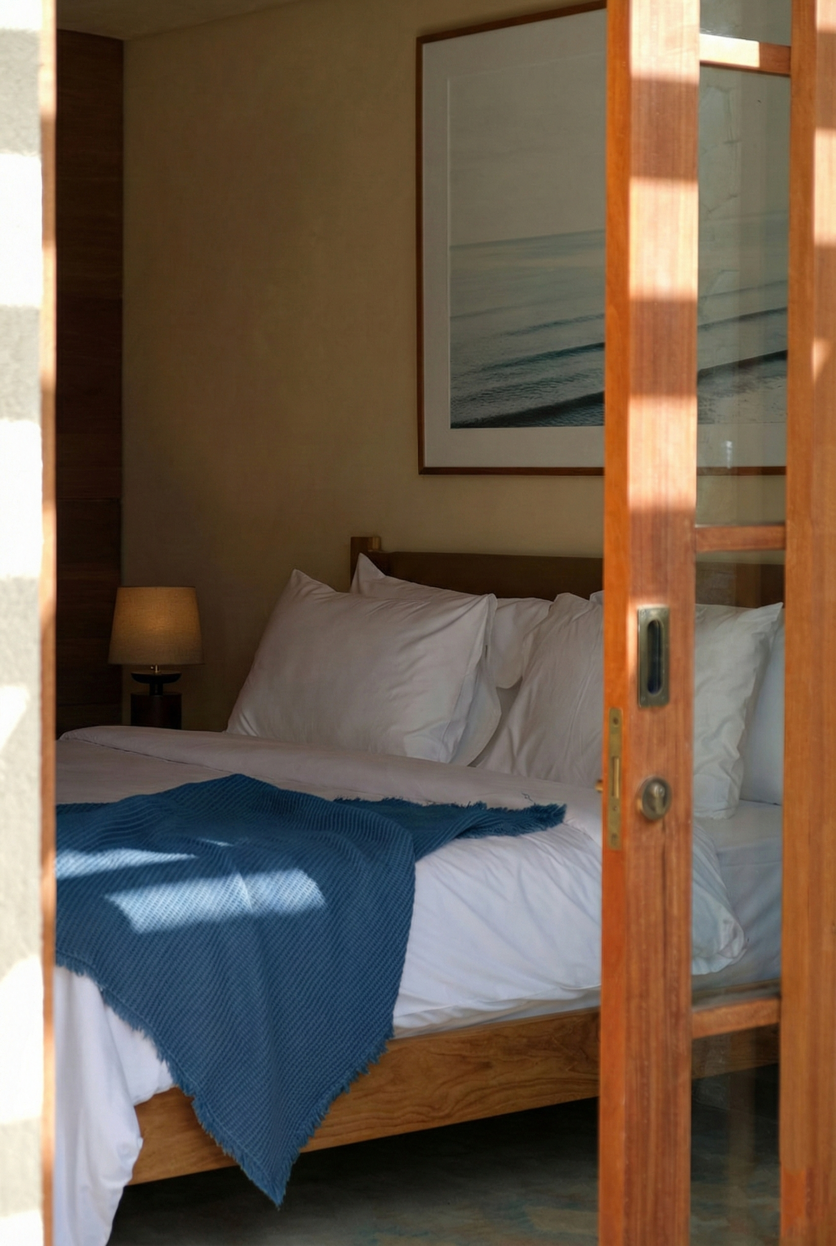 View of a bedroom with a bed covered in white sheets and pillows, a blue throw blanket, a framed ocean picture on the wall, a wooden nightstand with a lamp, and a partially opened wooden door with a glass panel.