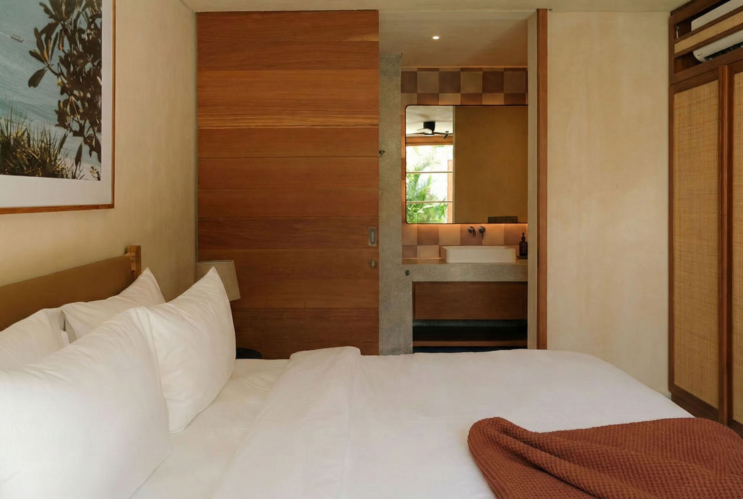A bedroom with a large bed covered in white linens, a wooden headboard, a bedside lamp, artwork on the wall, and a view of greenery through the window in the background.
