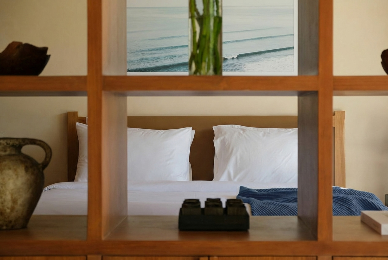 View of a bedroom through a wooden shelf, showing a bed with white pillows and a blue blanket, with a landscape painting of ocean waves hanging above.