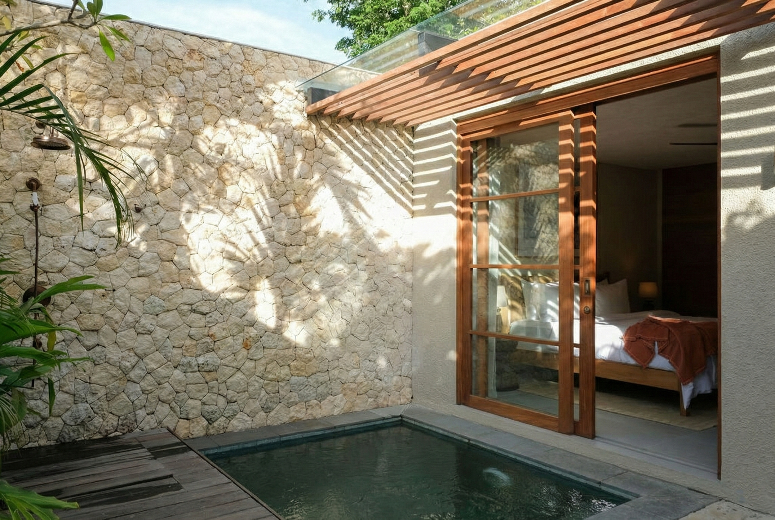 Exterior view of a modern house with a stone wall, wooden and glass sliding door, and a small pool or water feature with dark tiles, part of a patio with wooden decking, and greenery including a palm plant.