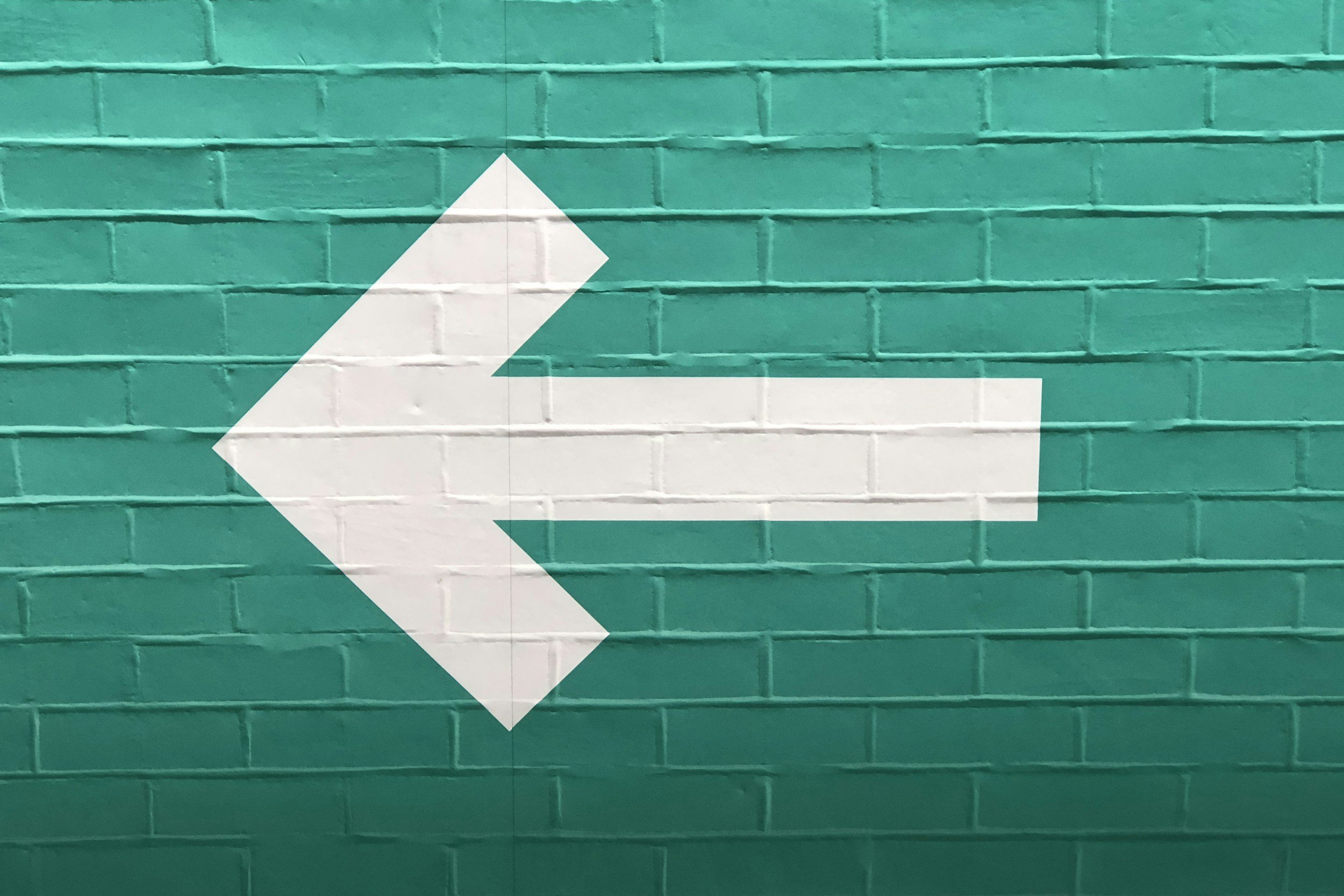 White left arrow painted on a teal brick wall.