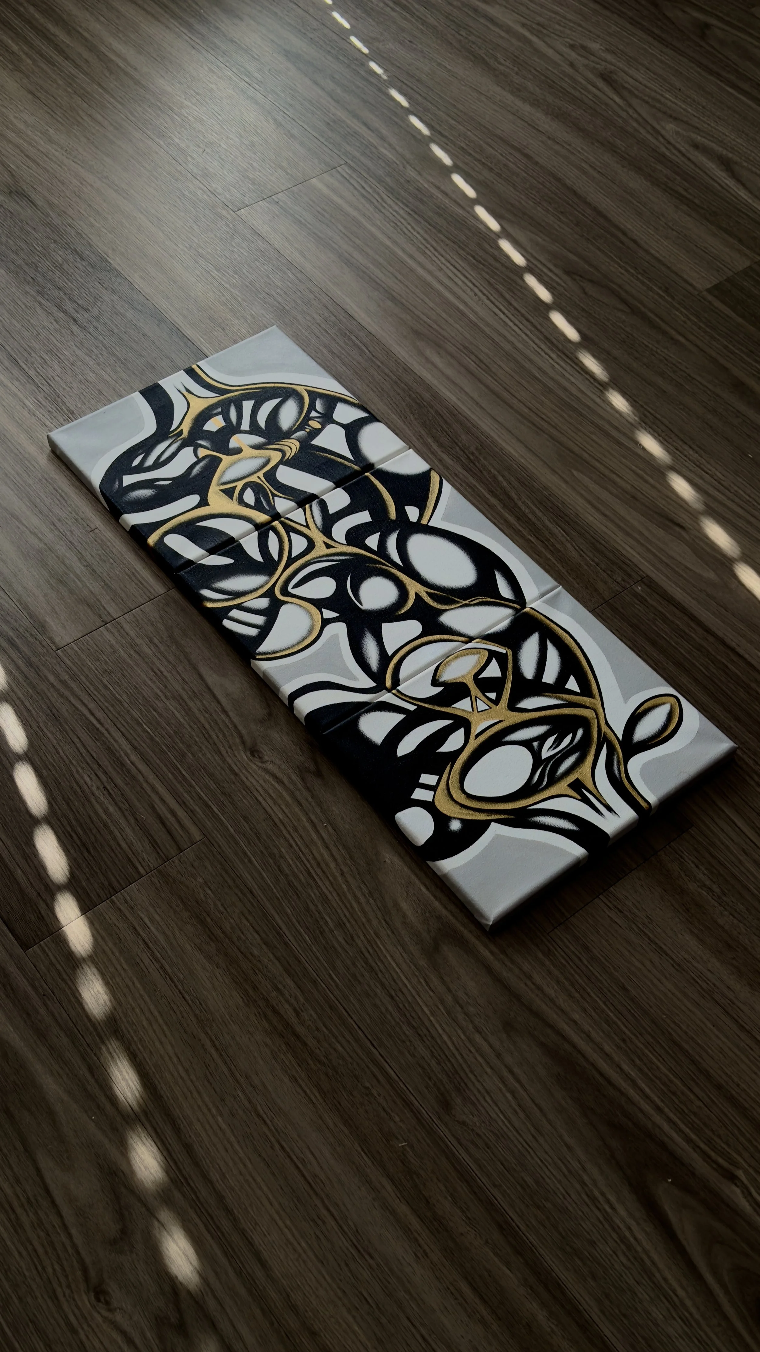 Abstract painting with black, white, and gold swirling lines on a rectangular canvas placed on a wooden floor.