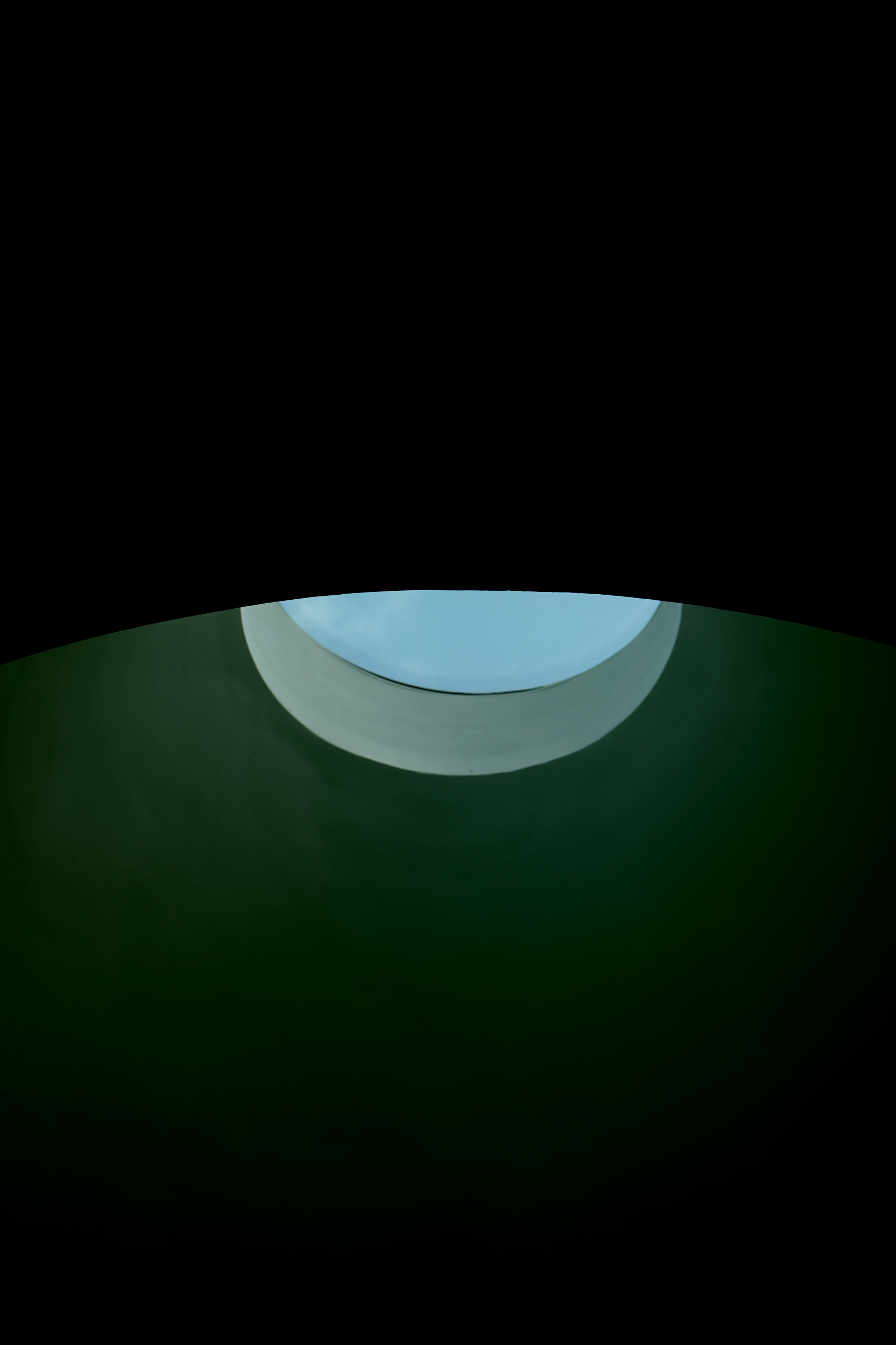 View looking up a green and white pipe with a circular opening showing a blue sky and clouds above.