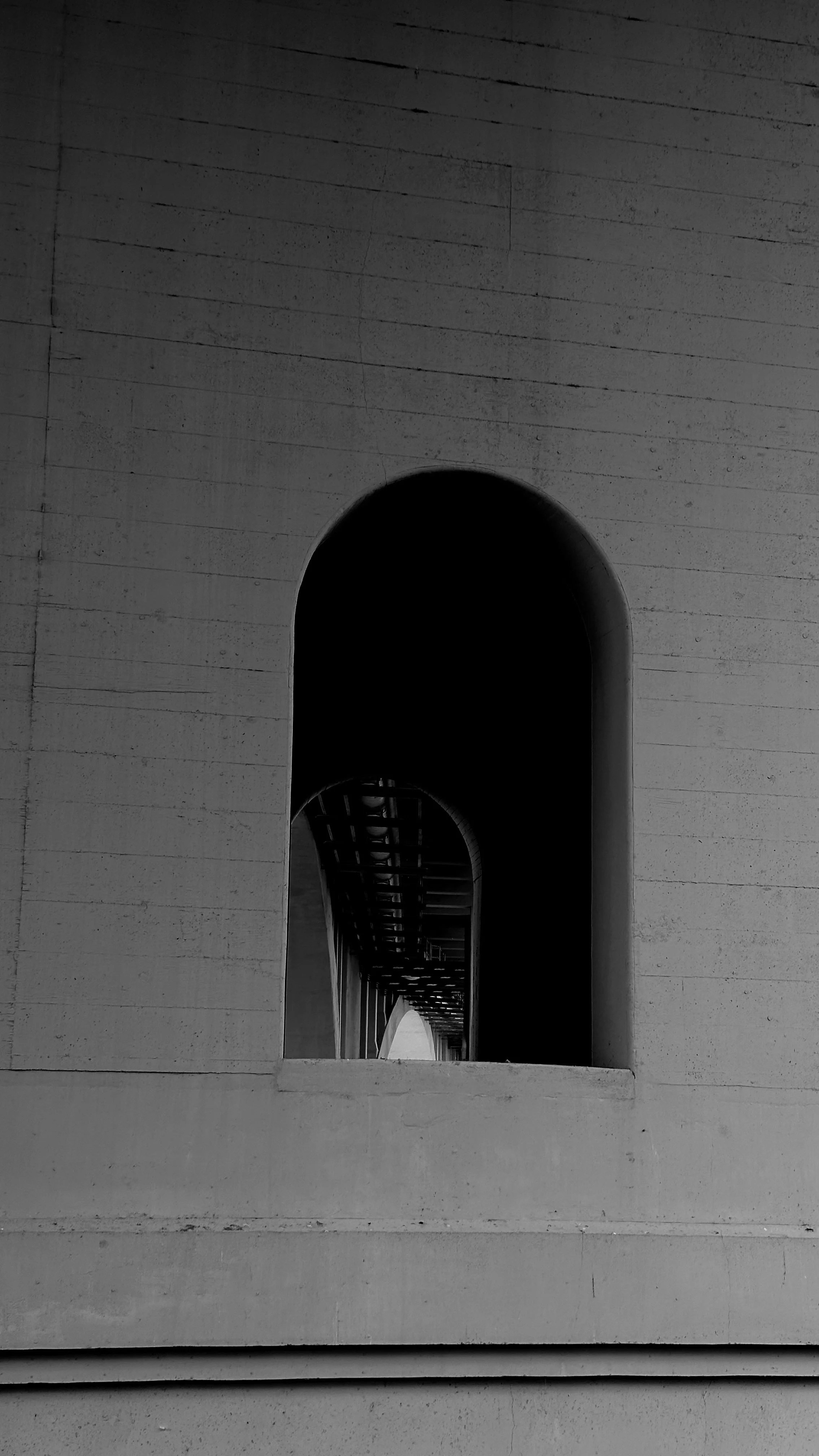 Black and white photo of a series of arched openings on a building, with a view of a bridge or walkway extending into the distance through the arches.