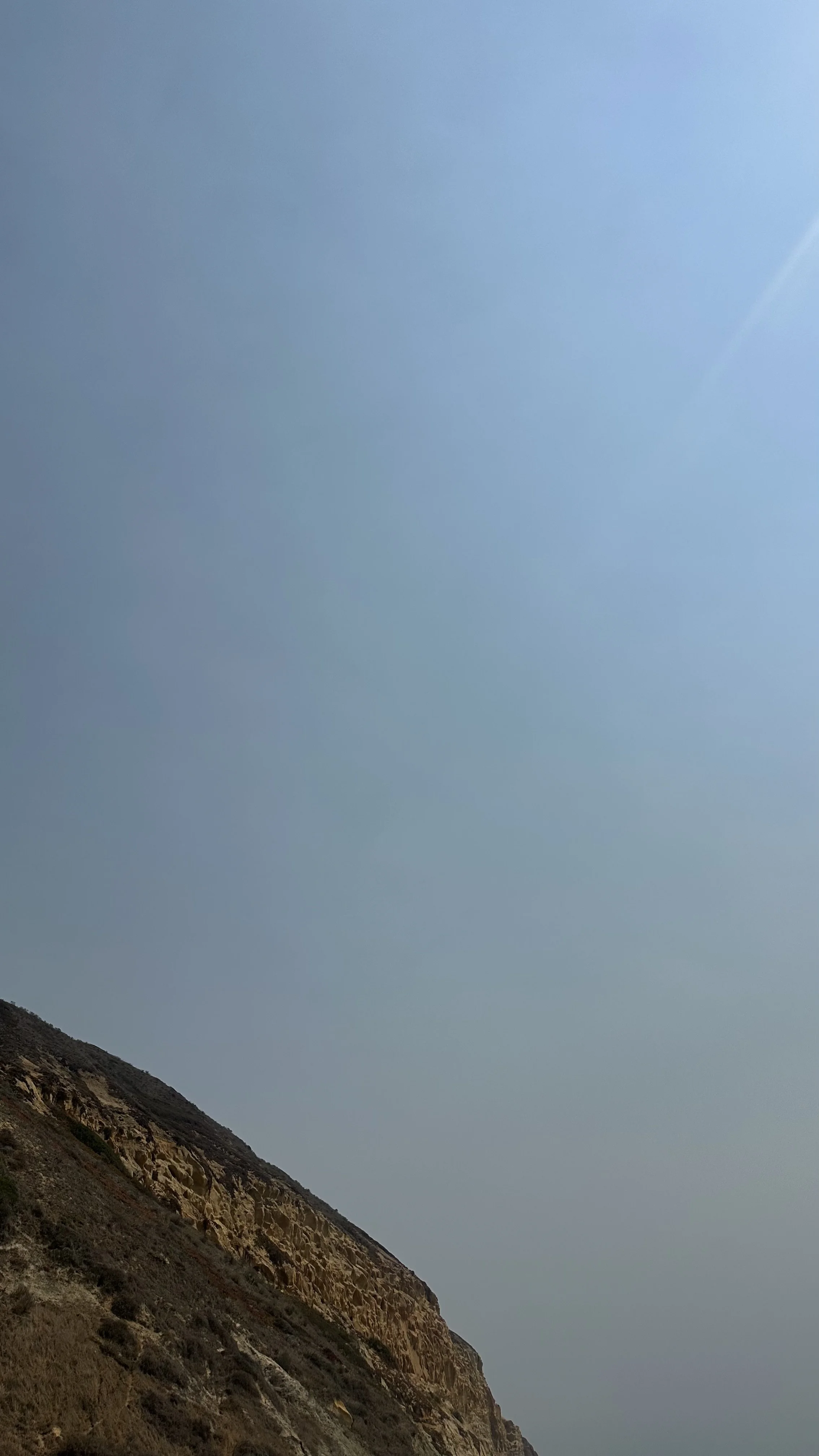 Part of a rocky hillside with sparse vegetation against a light blue sky.