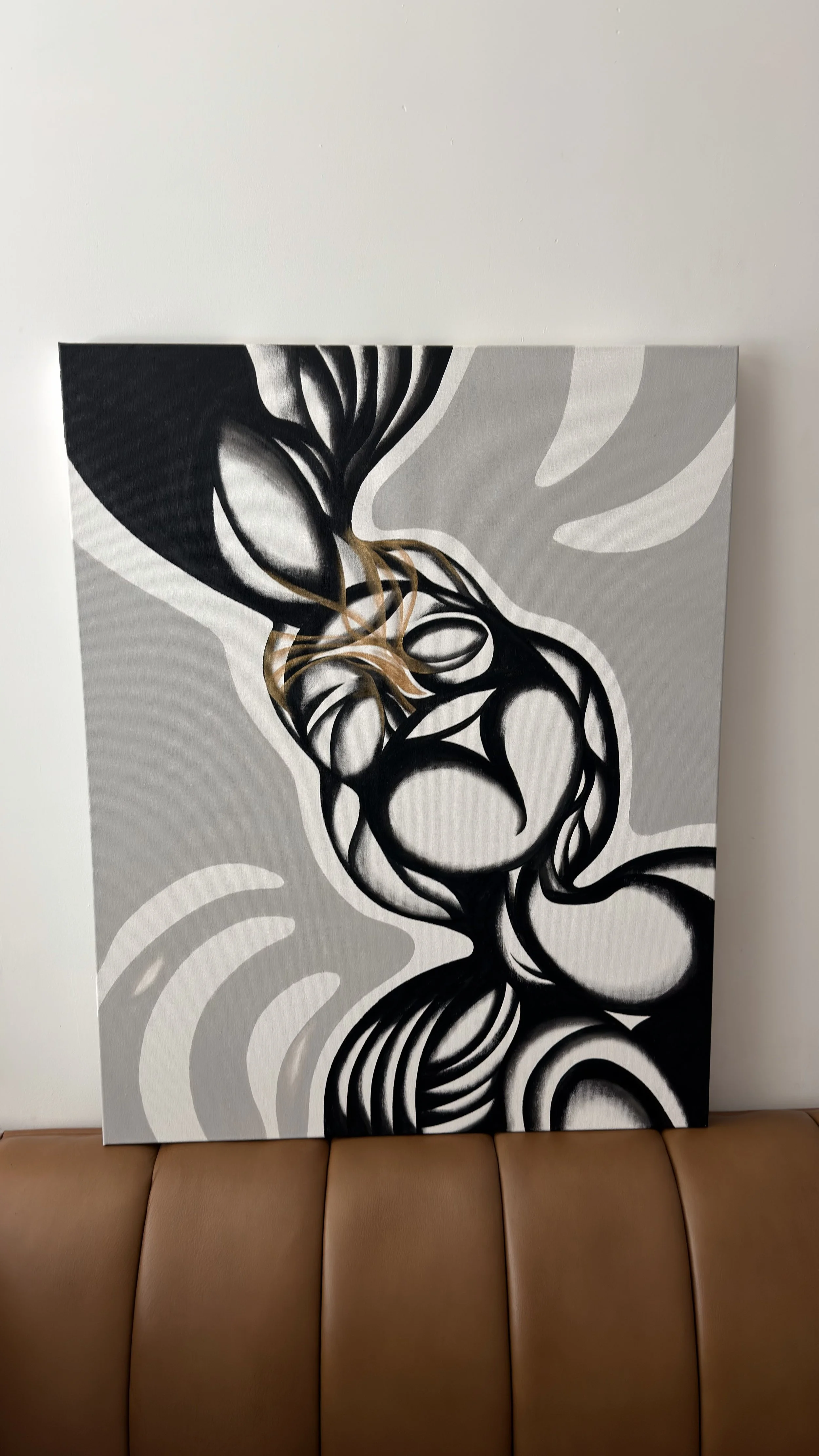 Abstract black and white artwork with swirling shapes and a hint of gold, leaning against a white wall on a brown leather couch.