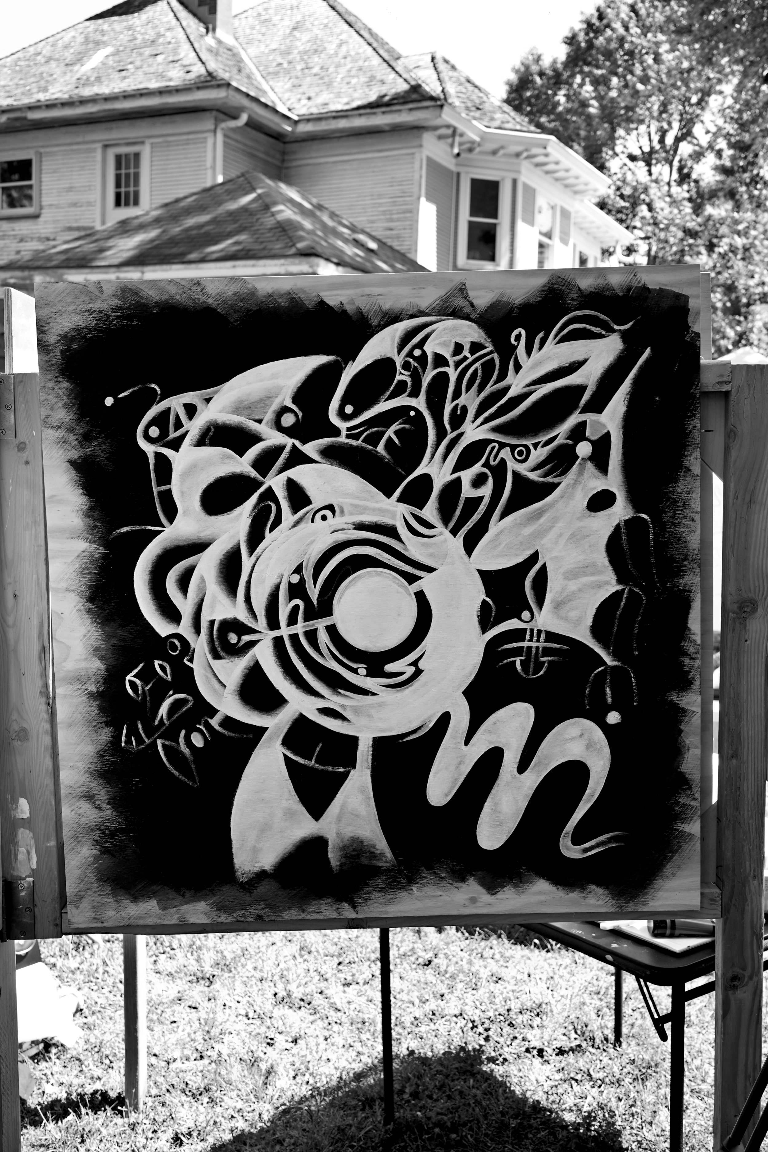 Black and white outdoor art display with abstract, flowing designs and lettering on a canvas, set up on a wooden frame in a yard with a house and trees in the background.