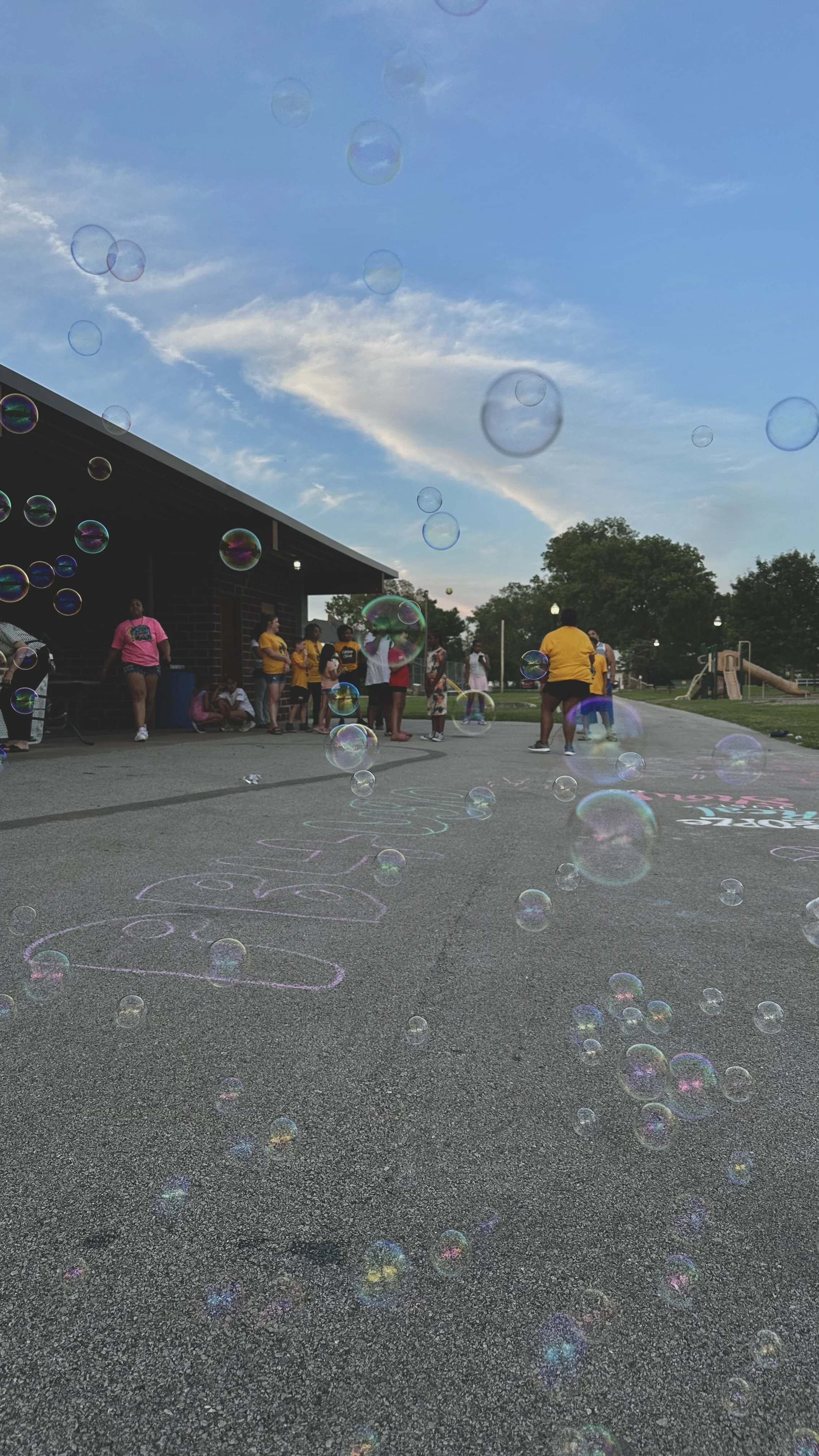 Children and adults gather outdoors around a building on a pavement with chalk drawings, with soap bubbles floating in the sky during daytime.