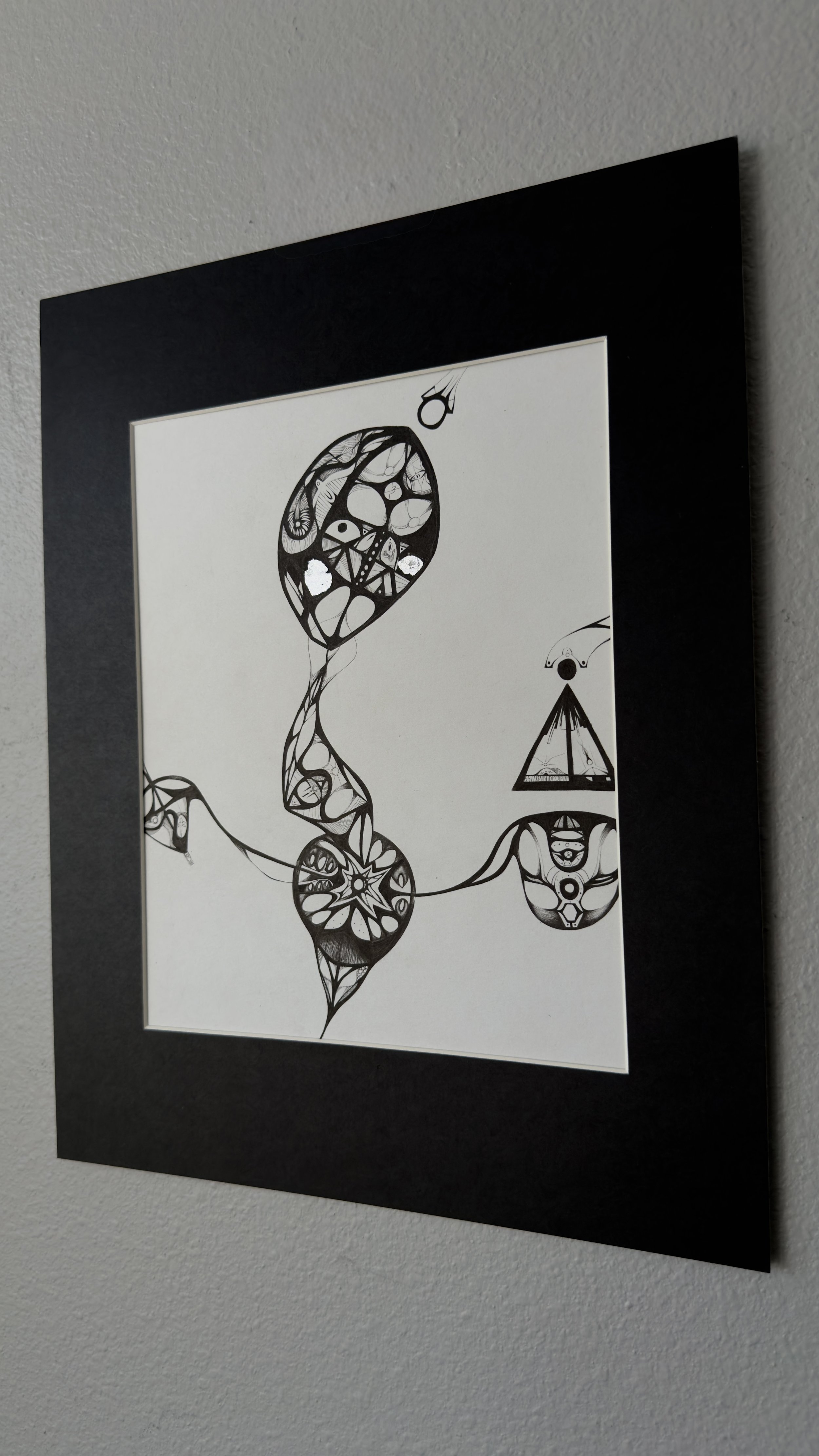 Black and white abstract art with interconnected shapes and patterns, including circles, triangles, and lines, framed in a black border on a white wall.