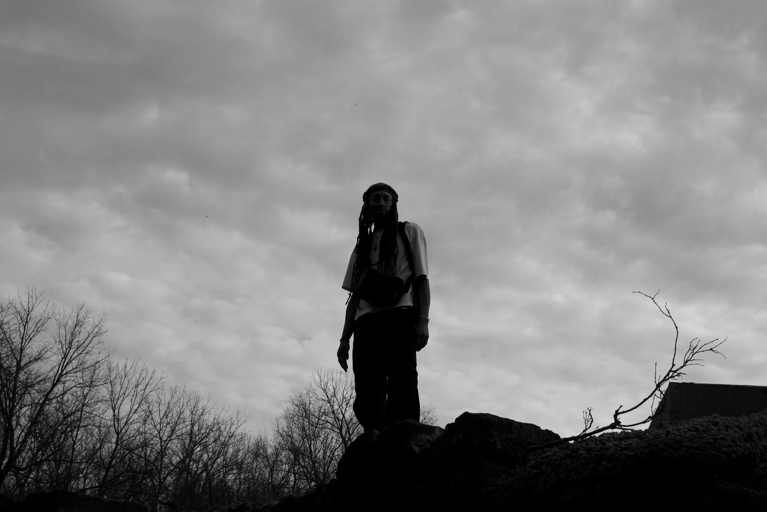 A non-binary individual with locs stands on a rock against a cloudy sky, outdoors, with leafless trees and a branch in the foreground.