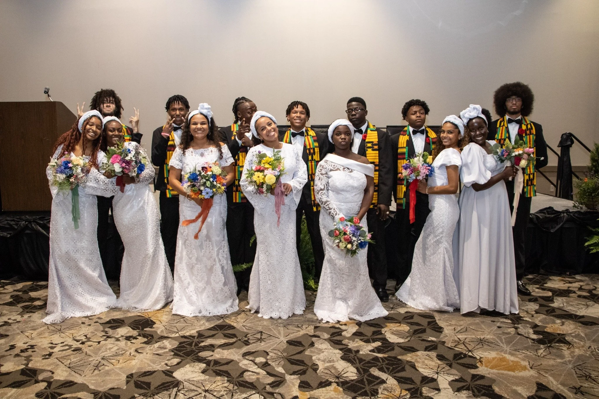 A group of young people dressed in formal attire, some in white dresses with floral bouquets, and others in black suits with colorful Kente cloth scarves, standing together on a patterned carpet in an indoor setting.