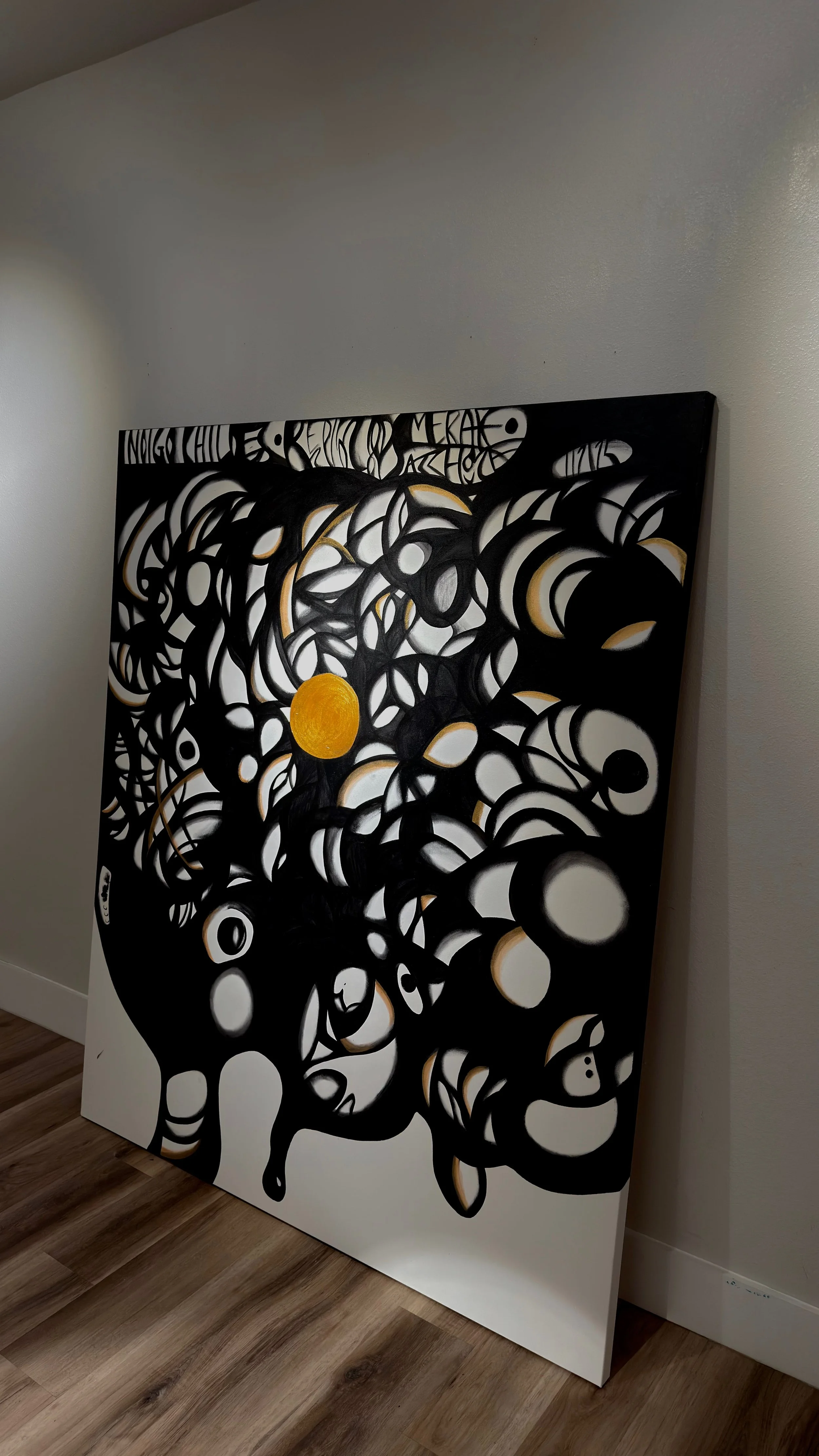 A black and white abstract painting featuring swirling lines and circles, with some gold accents, leaning against a white wall in a room with wooden floors.