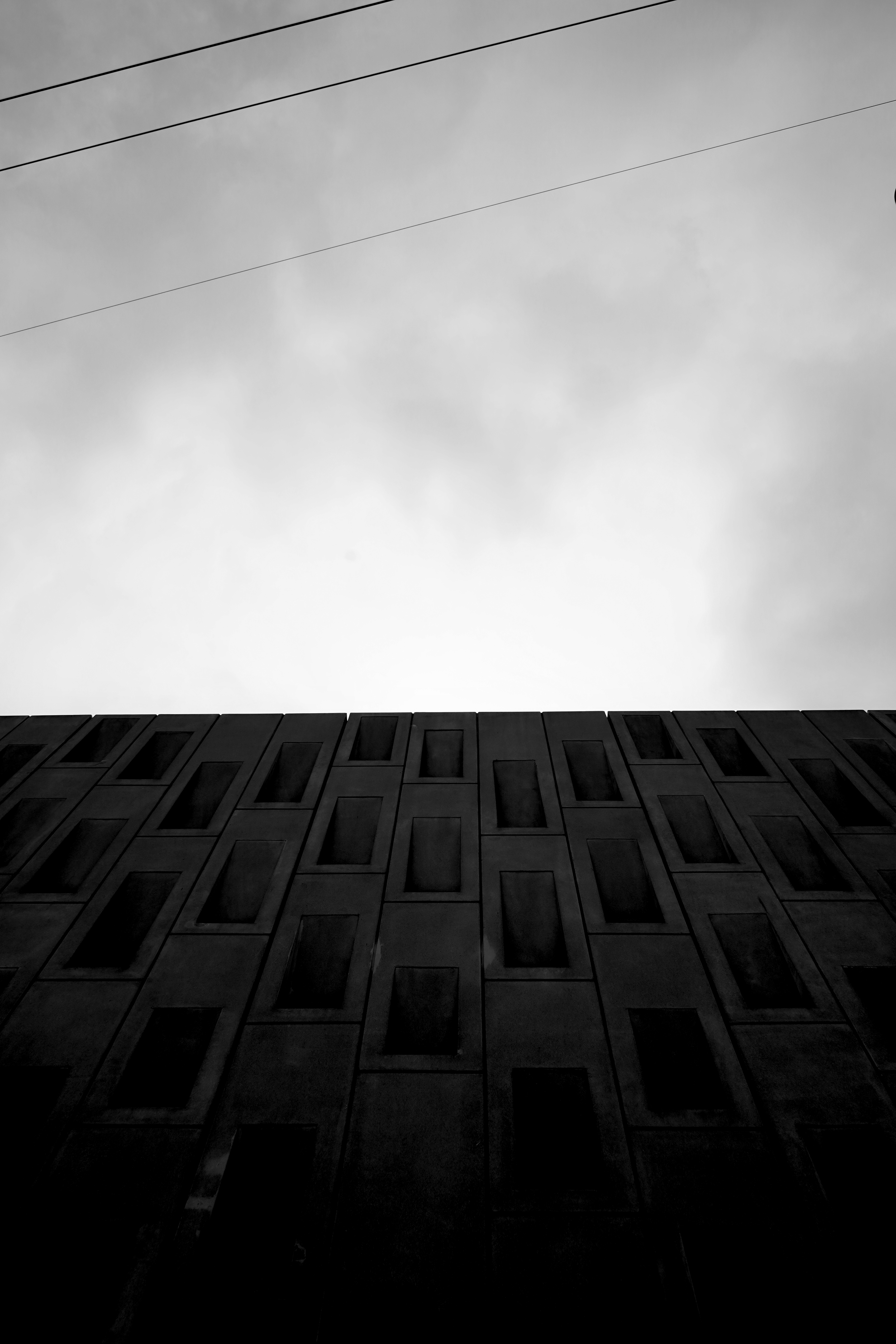 Low-angle view of a modern building with geometric windows against a cloudy sky. Several power lines are visible overhead.