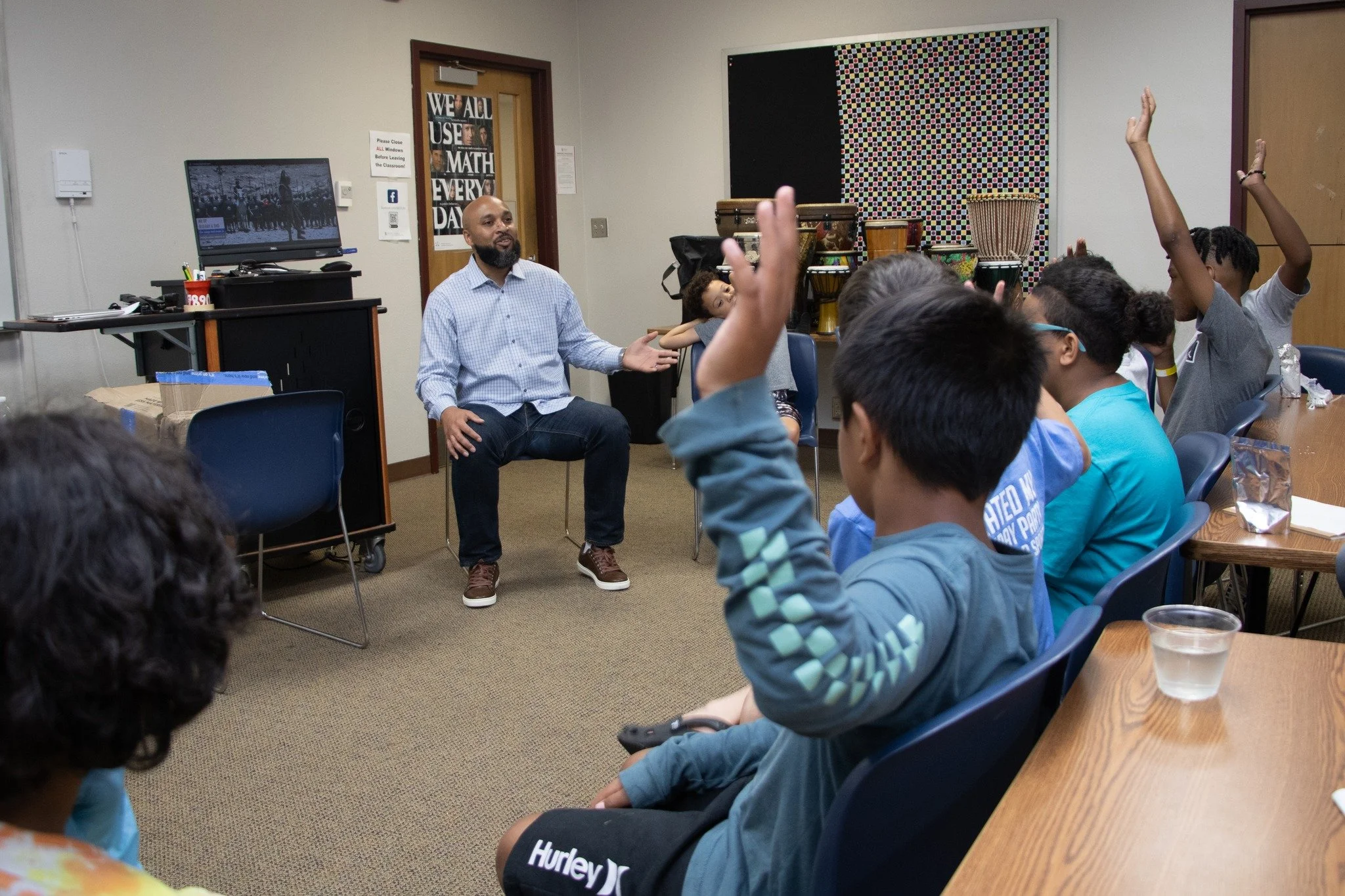 A teacher or presenter talking to a group of students in a classroom setting, with some students raising their hands.