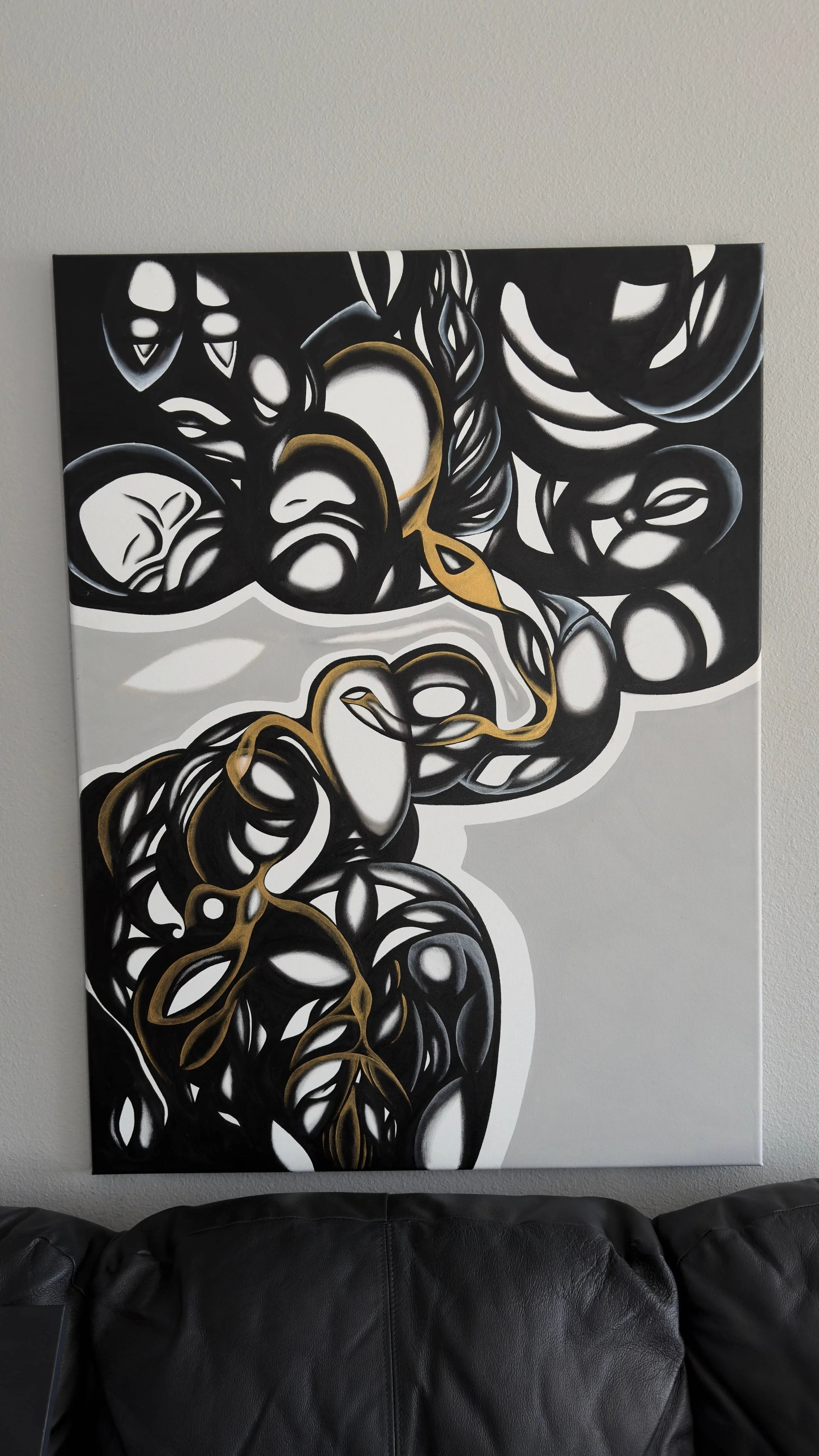Abstract black, white, and gold painting on canvas hanging on a wall, with part of a black leather couch visible at the bottom.