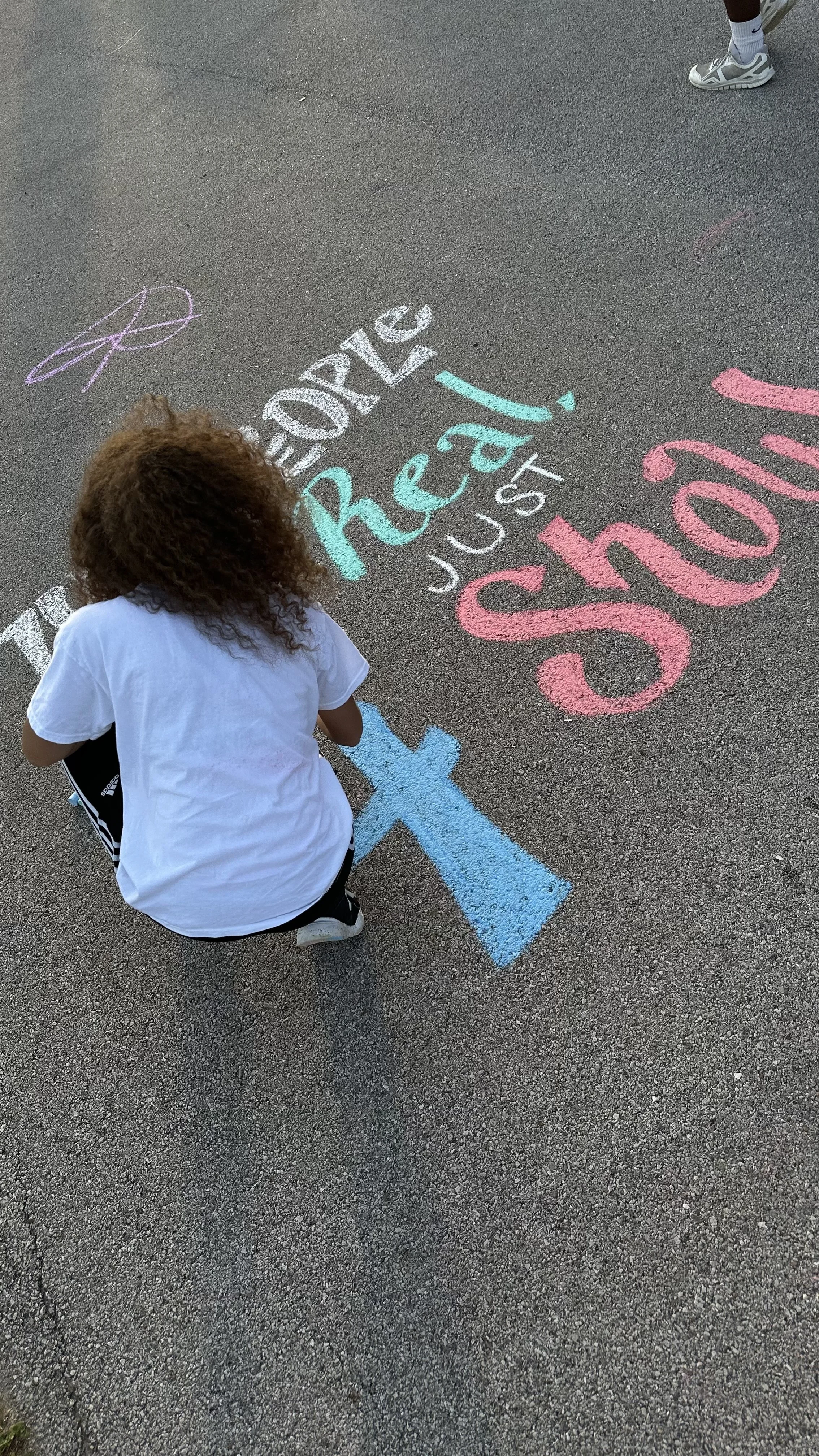 Child with curly hair writing on the pavement with colorful chalk that says "Please read just one story."