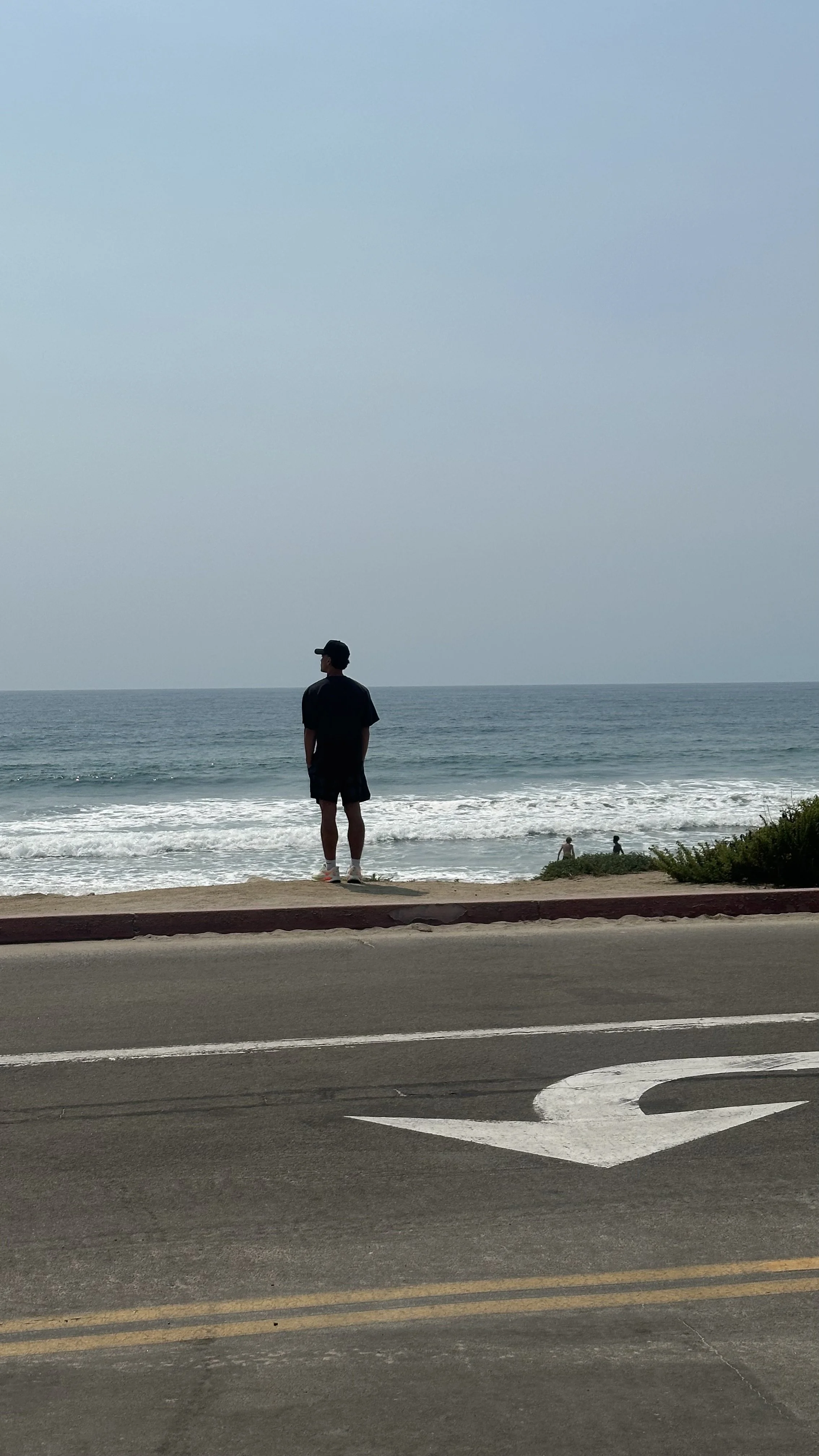 A person wearing black shorts, a black t-shirt, and a black cap standing on the sidewalk, looking at the ocean and waves, with two people in the water.