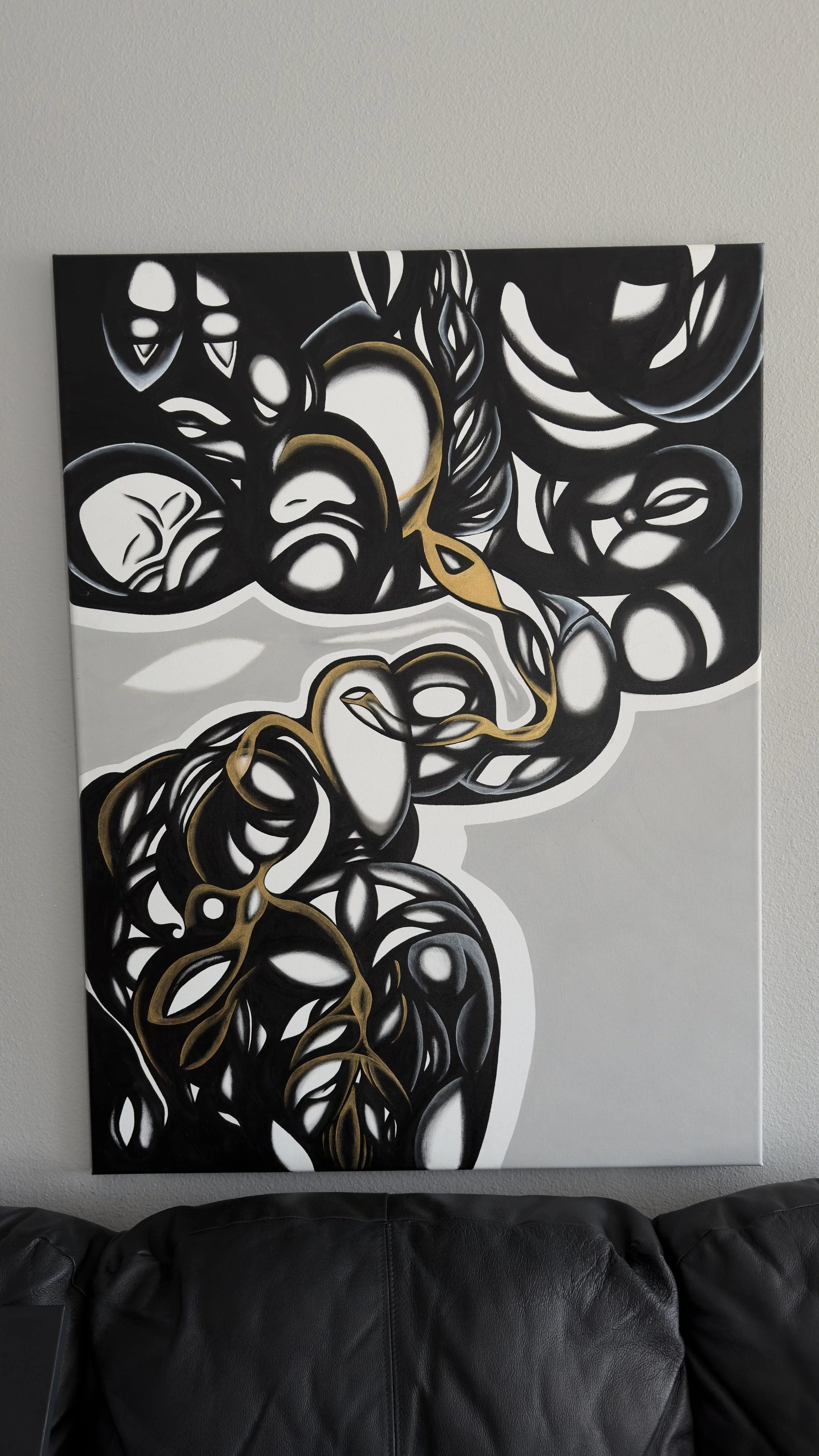 Abstract black, white, gray, and gold artwork featuring flowing, intertwined shapes and faces, mounted on a wall above a black leather couch.
