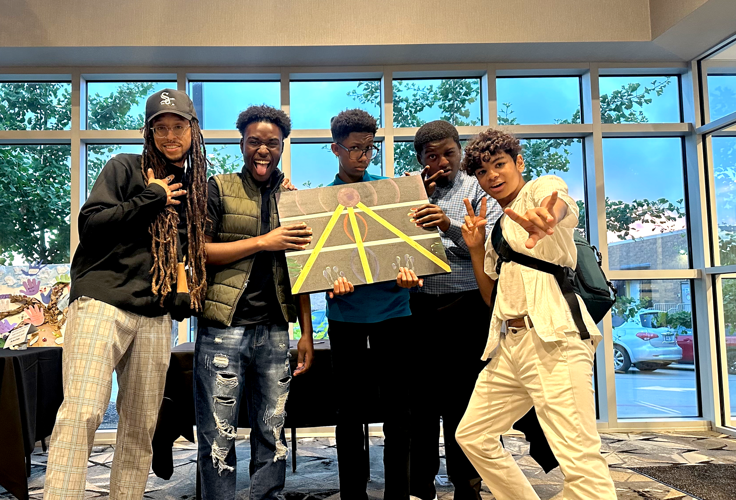 Five young men standing indoors in front of large windows, holding a painted canvas with yellow lines and a purple circle, with trees and parked cars visible outside.