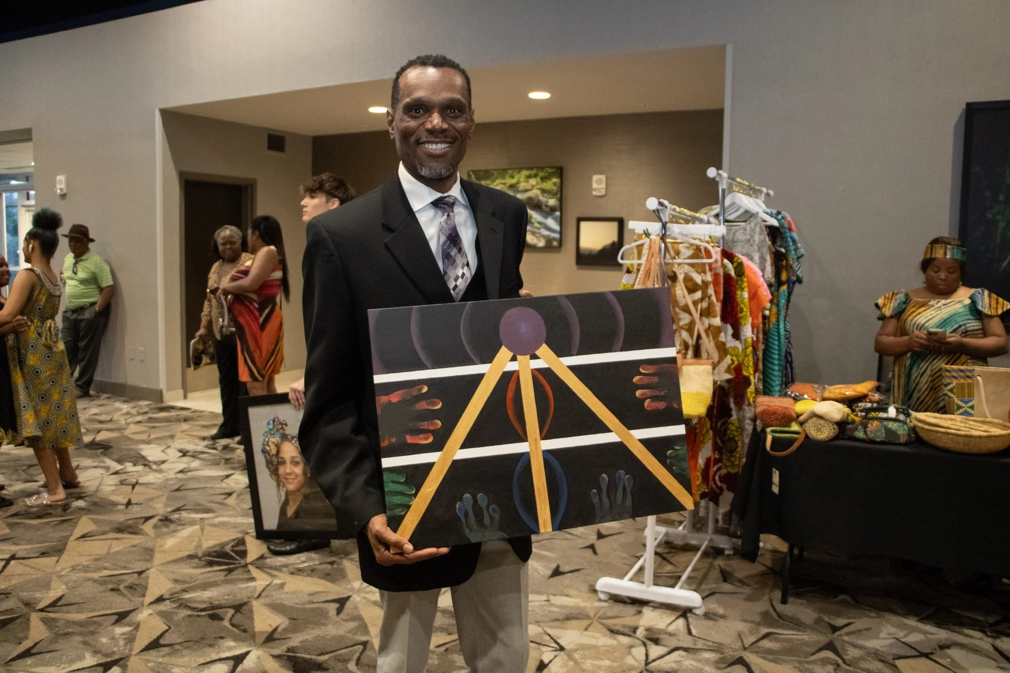 A smiling man in a suit holding an abstract painting at an indoor event with other people and display tables.