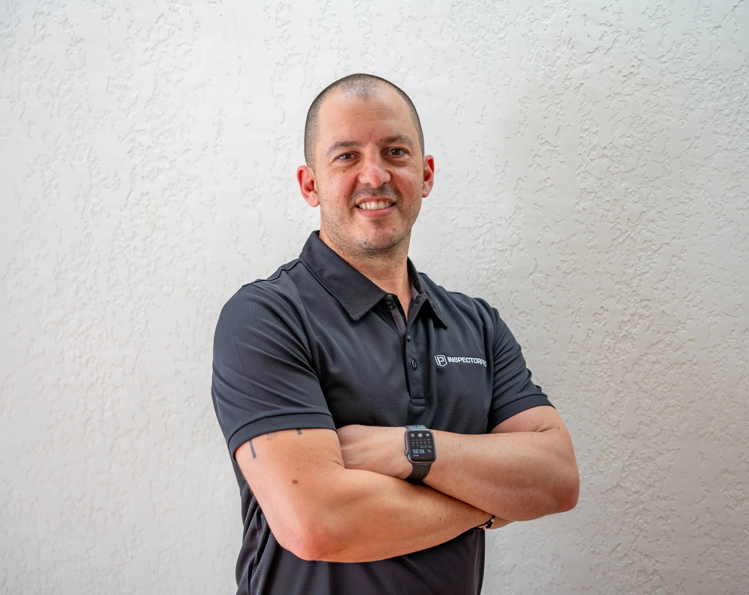 A man with a shaved head wearing a black polo shirt with logo, standing with arms crossed against a beige textured wall.