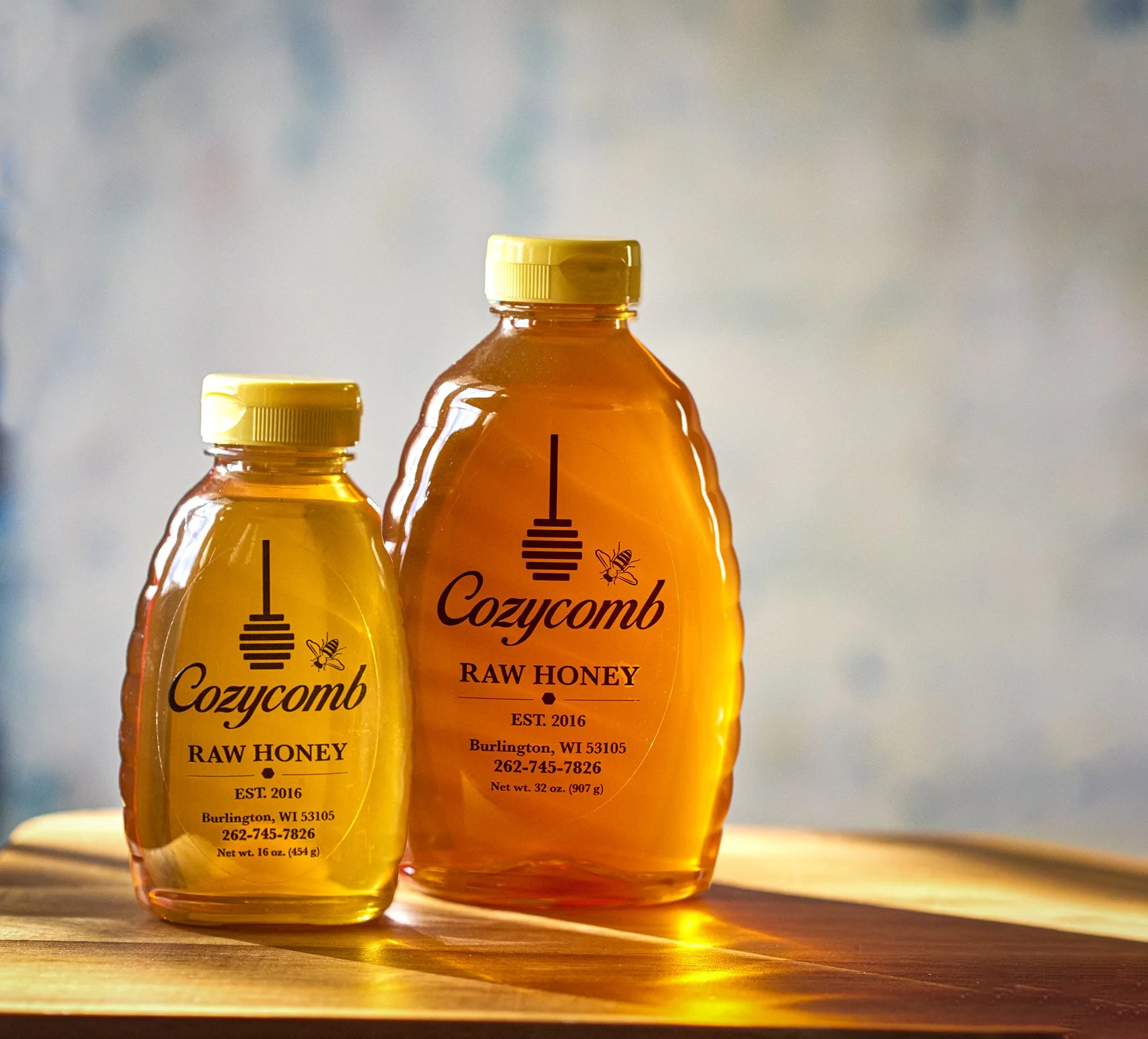 Two amber-colored bottles of Cozycamb raw honey with yellow caps on a wooden surface, sunlight shining through, with a blurred background.