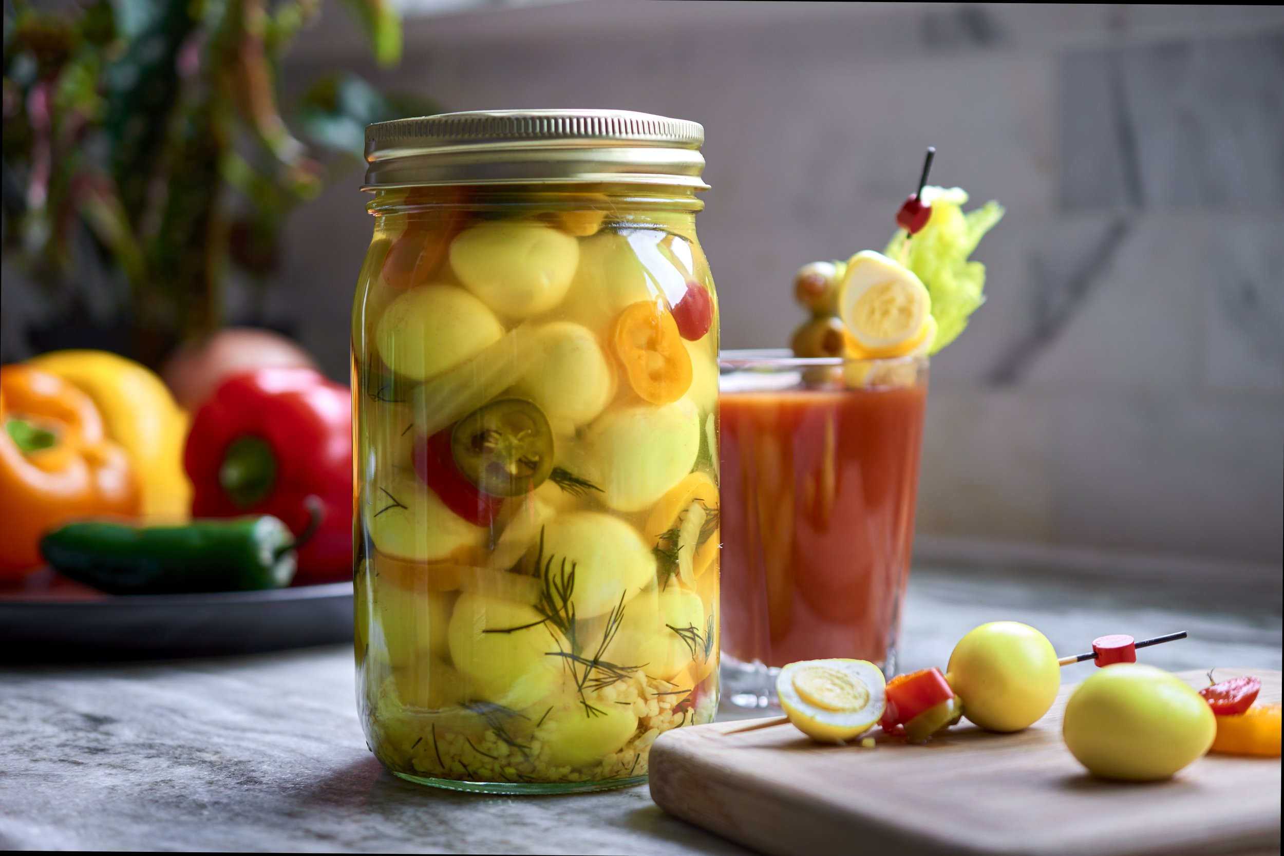 A glass jar filled with pickled vegetables, including yellow cucumbers, orange carrots, and green peppers, on a kitchen countertop with a cutting board and chopped vegetables nearby.