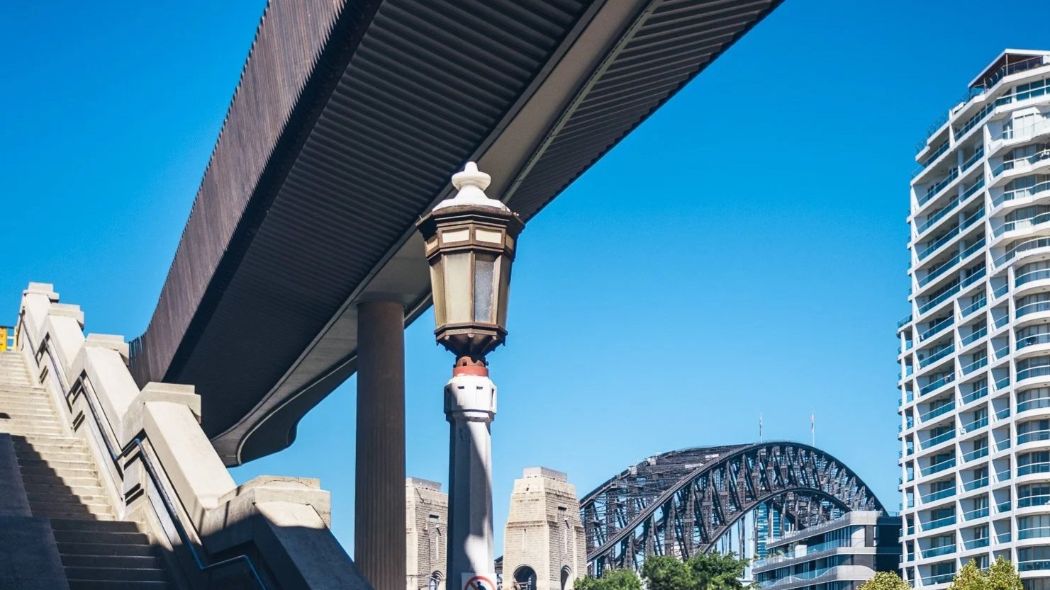 Street view in Sydney, Australia, showing the Harbour Bridge, modern buildings, and an overpass against a clear blue sky.