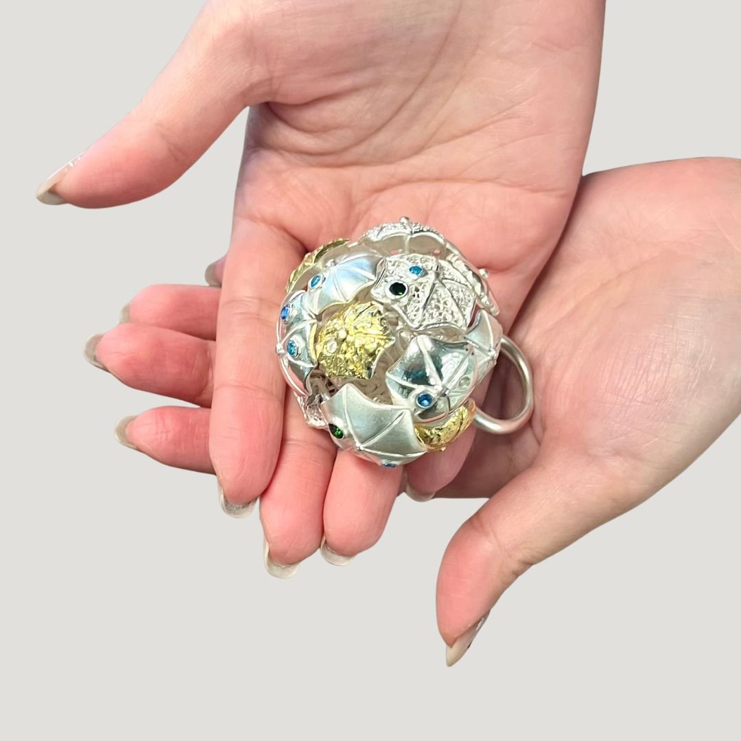 Close-up of a hand holding a spherical, silver and gold jewelry piece with various small decorative elements and colored gemstones.