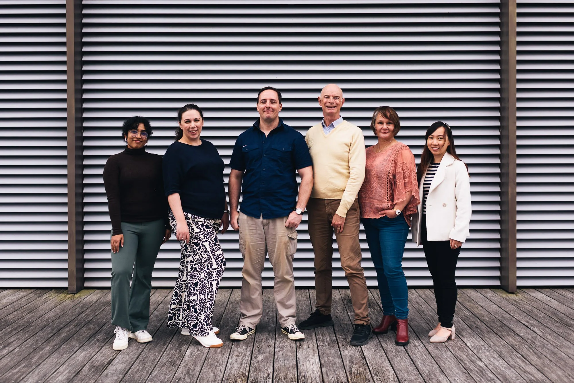 Group of six diverse people standing outdoors on a wooden deck in front of a metal slatted wall, smiling at the camera.