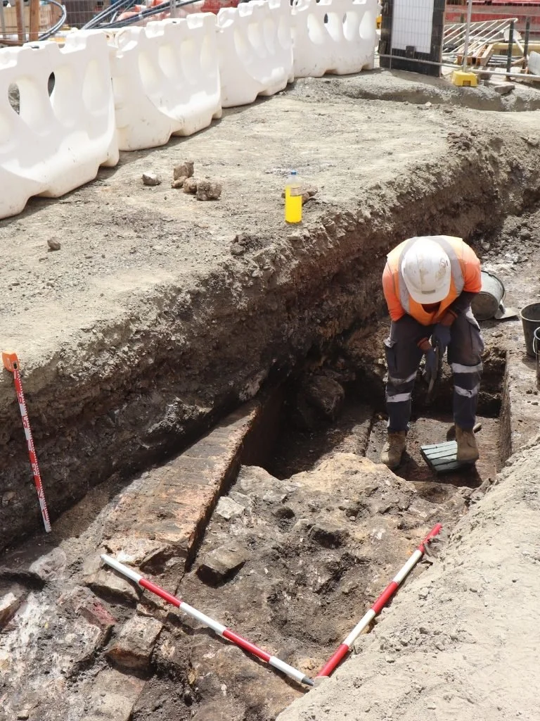 Construction worker inspecting an excavation site with exposed brick and soil, wearing a hard hat and safety gear, surrounded by construction barriers and equipment.
