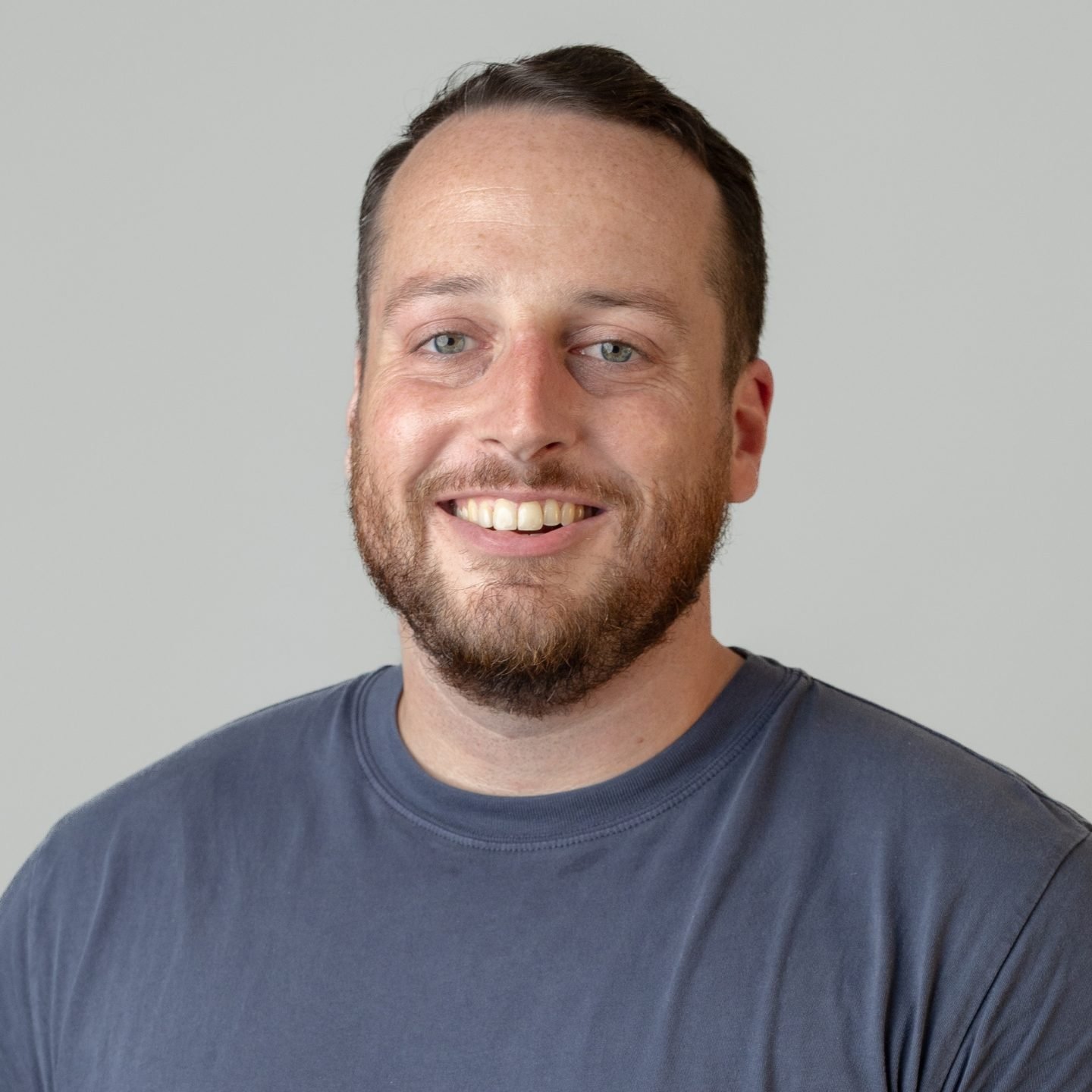 A smiling man with a beard and short hair wearing a gray t-shirt standing against a plain light gray background.