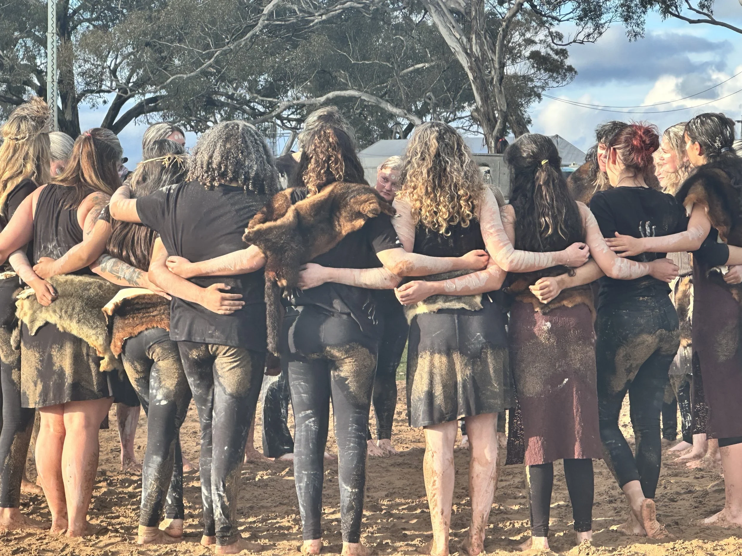 A group of women standing in a circle outdoors during daytime, embracing and holding each other, with trees and a cloudy sky in the background.
