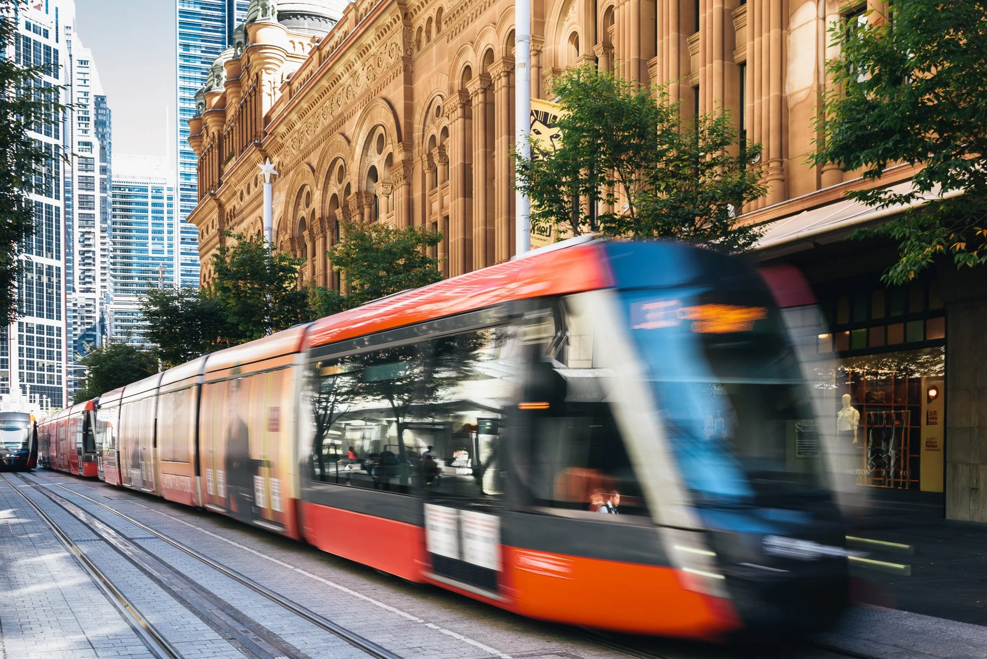 Red and white modern tram moving along tracks in a city street with tall buildings and trees.