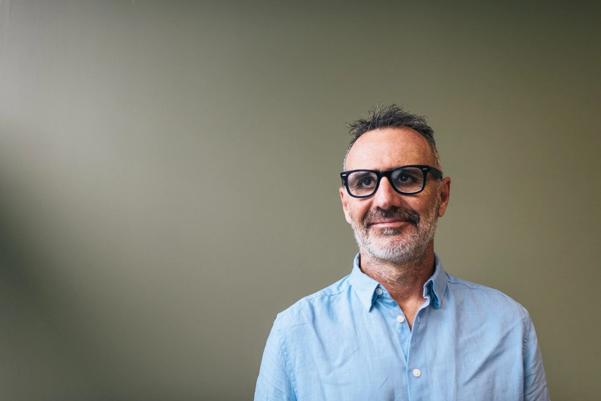 A middle-aged man with gray hair, glasses, and a beard, wearing a light blue button-up shirt, stands against a plain, muted wall.