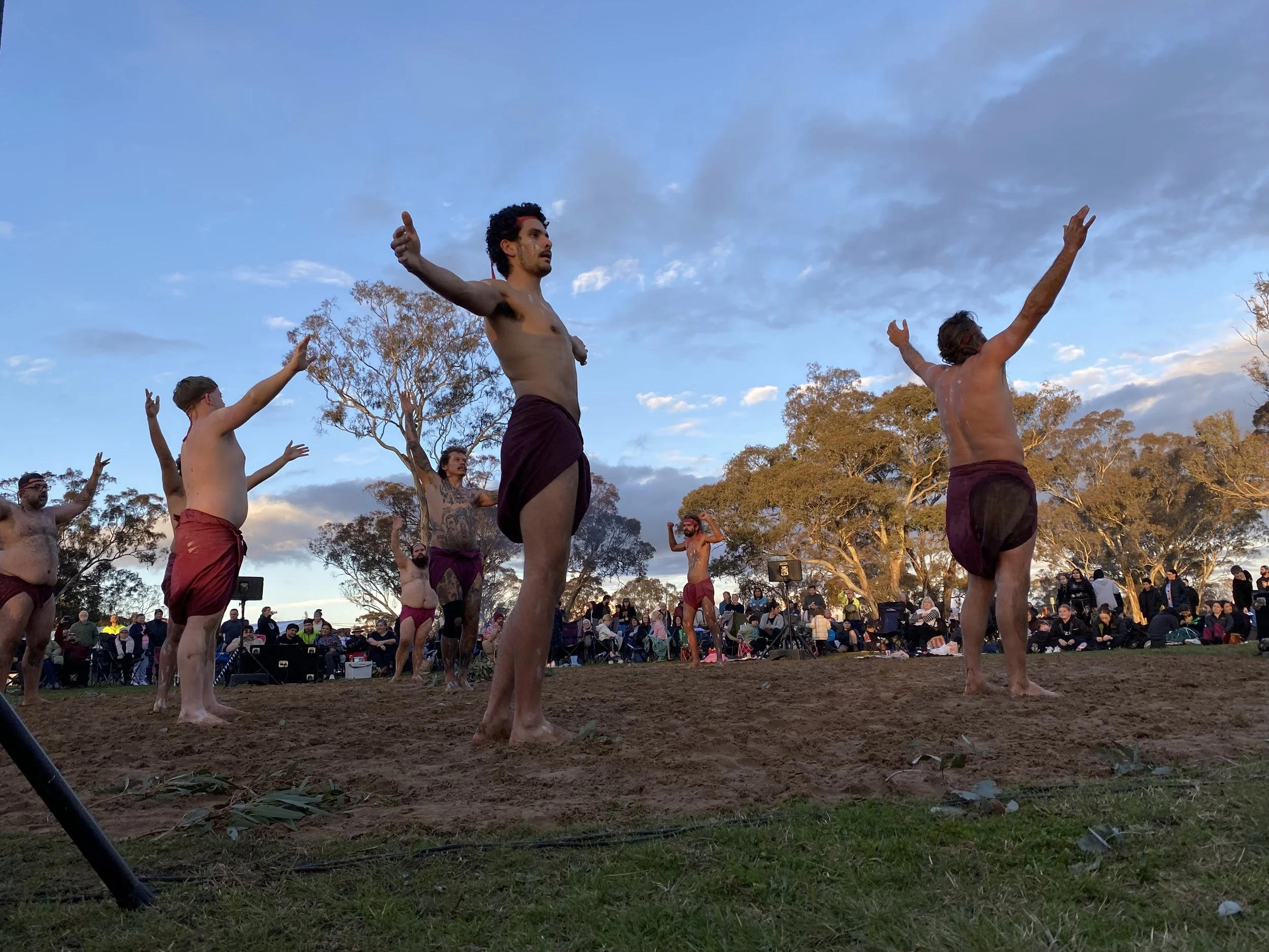 A group of men practicing a traditional dance or martial arts outdoors, with some men shirtless and others wearing dark shorts, raised arms, and focused expressions, gathered in front of a large audience seated on the grass under trees during sunset.