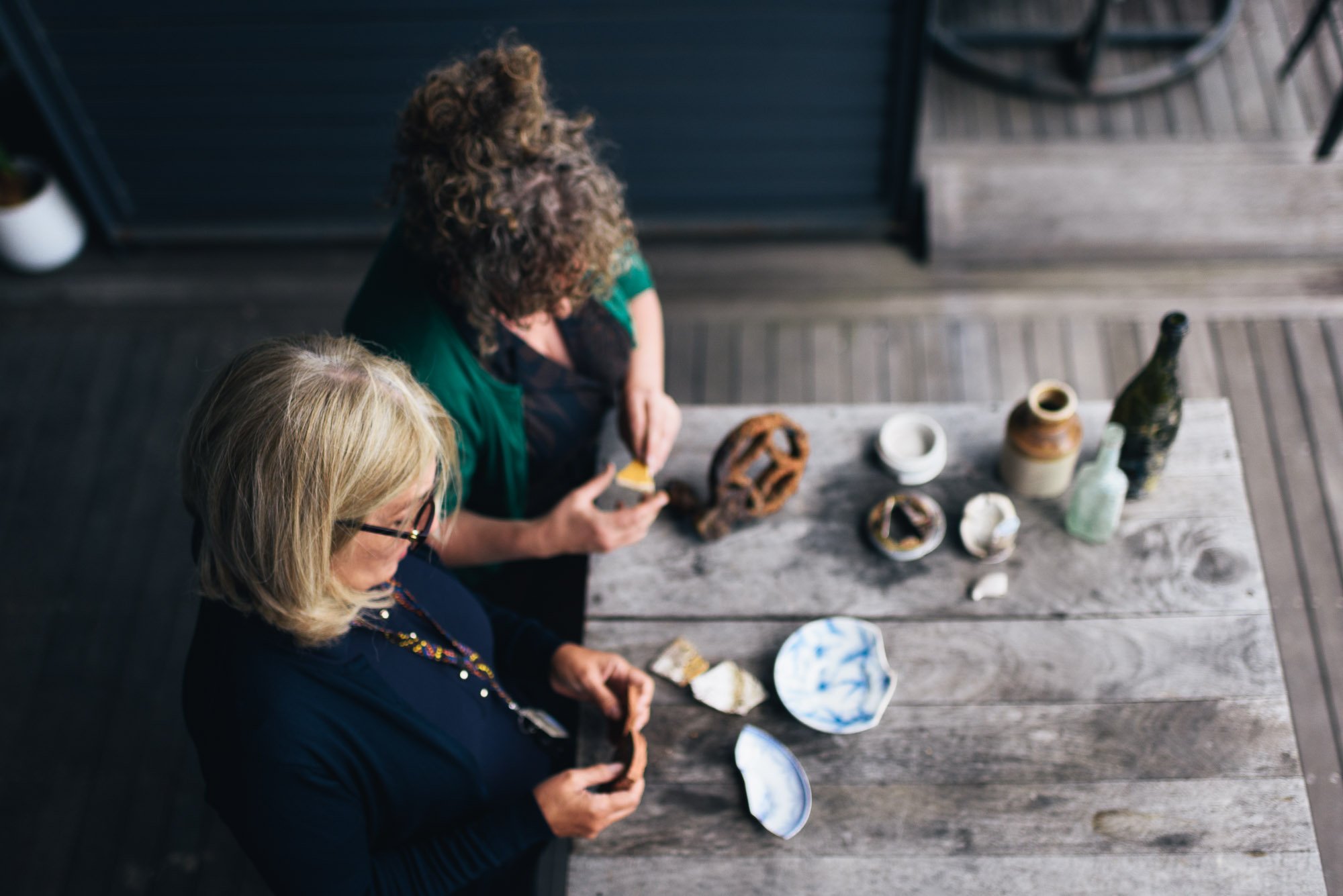 Two women sitting at a wooden table looking at pottery pieces and vases, with a dark deck and outdoor furniture in the background.