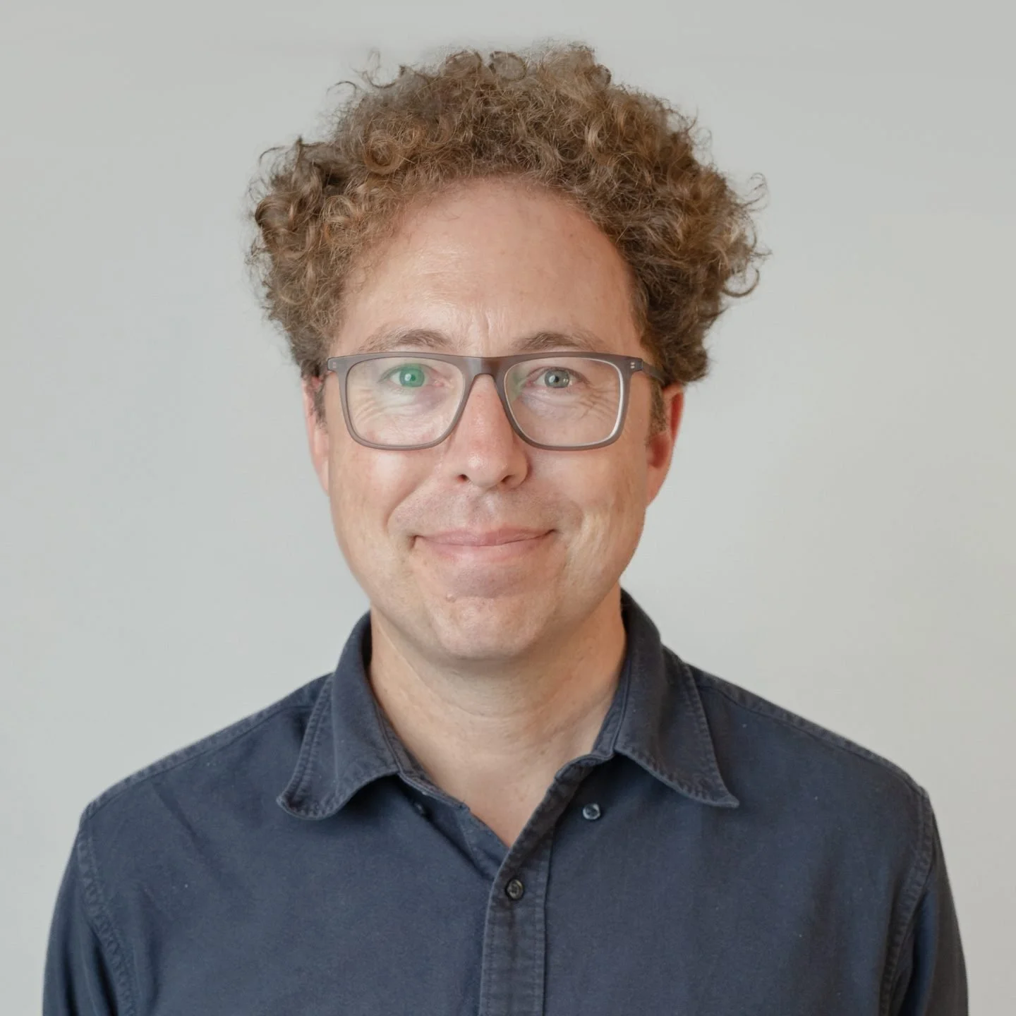 A man with curly brown hair and gray glasses, wearing a dark blue shirt, smiling at the camera against a plain light gray background.
