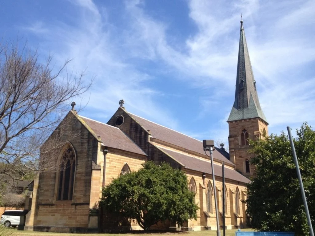 A traditional stone church with a tall steeple, surrounded by trees, under a blue sky with some clouds.