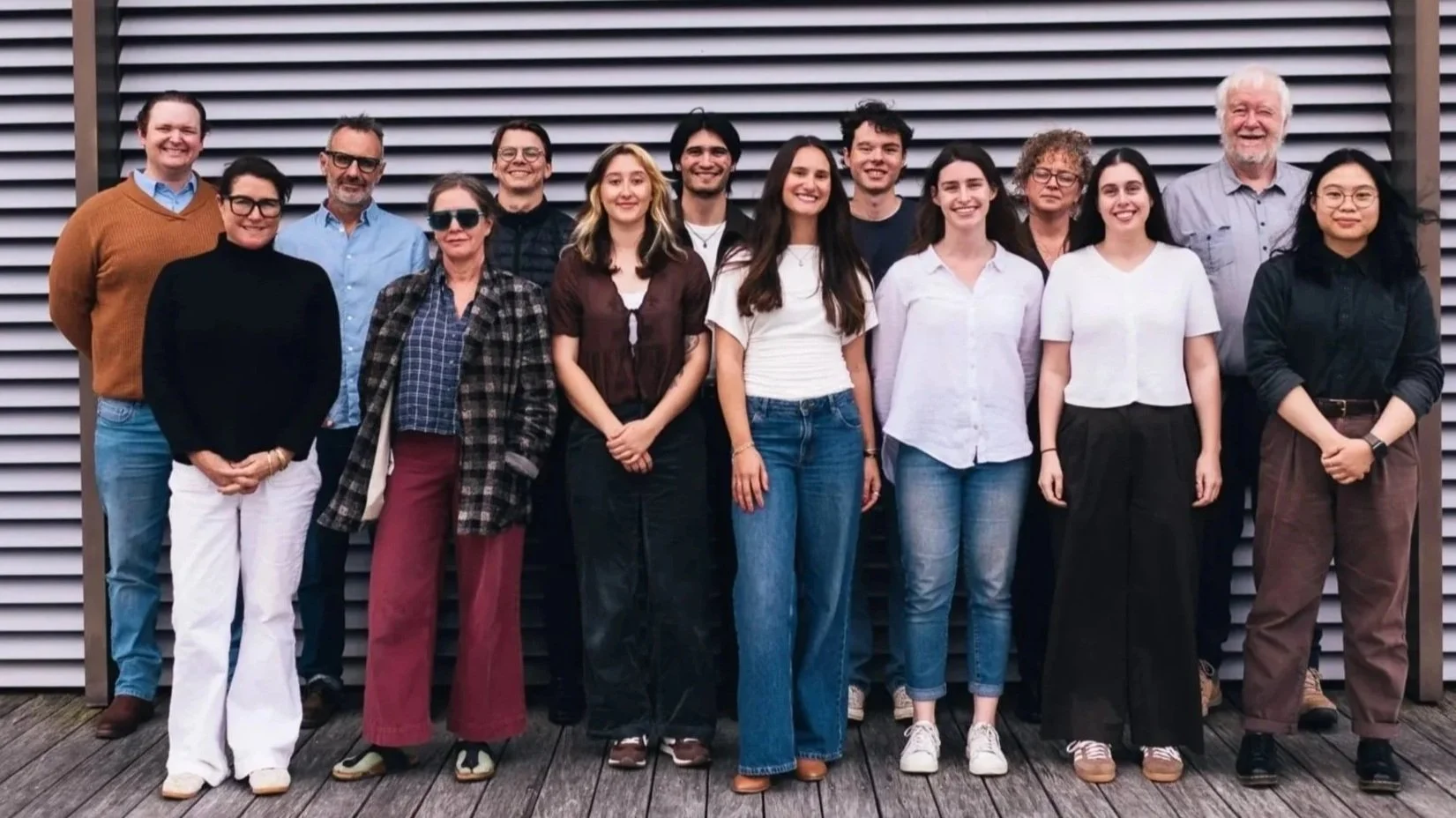 Group of fifteen diverse people standing outdoors in front of a metal slat wall, posing for a group photo.