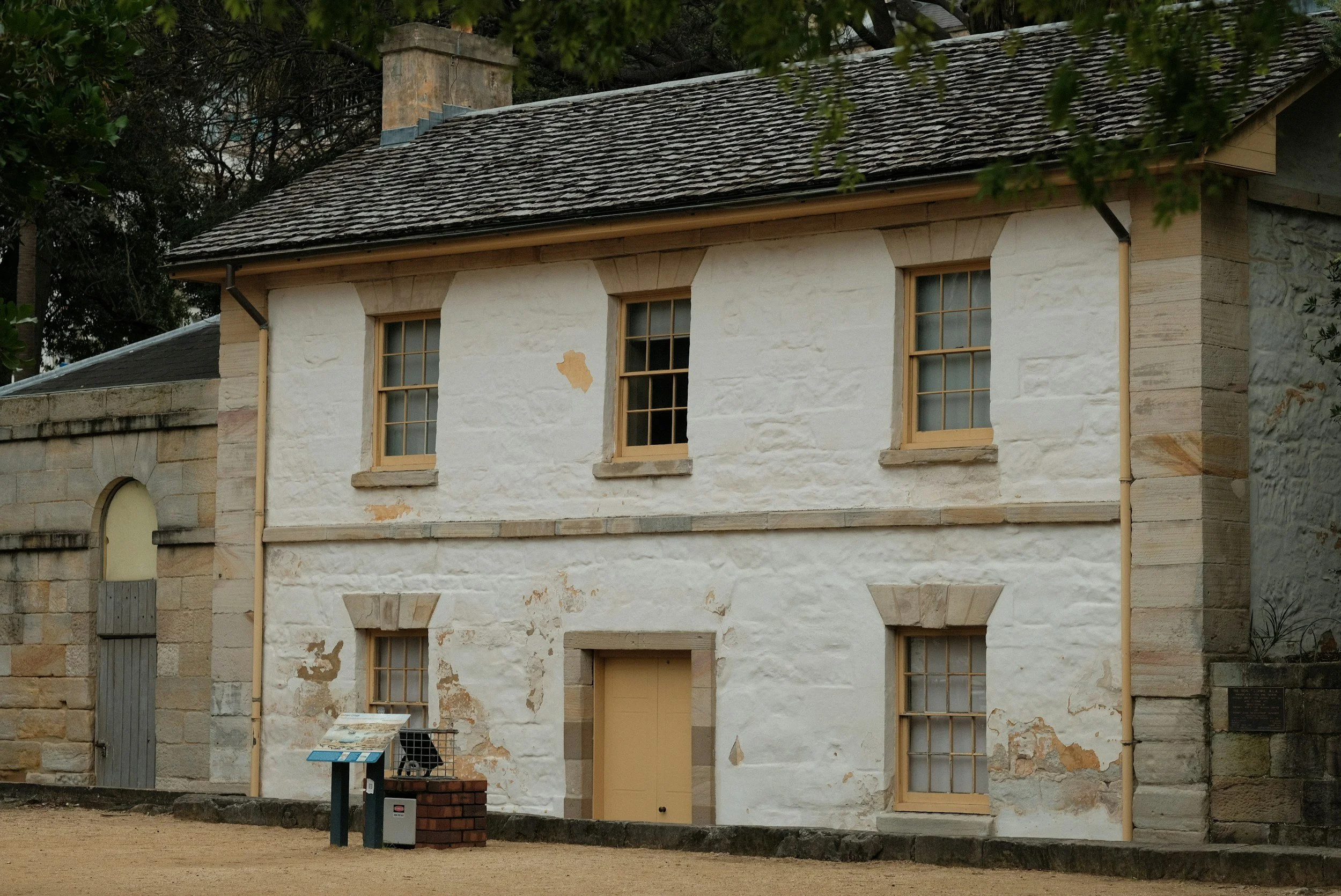Historic stone building with a white painted facade, wooden window frames, and a gabled roof with dark shingles, located in a park or outdoor setting.