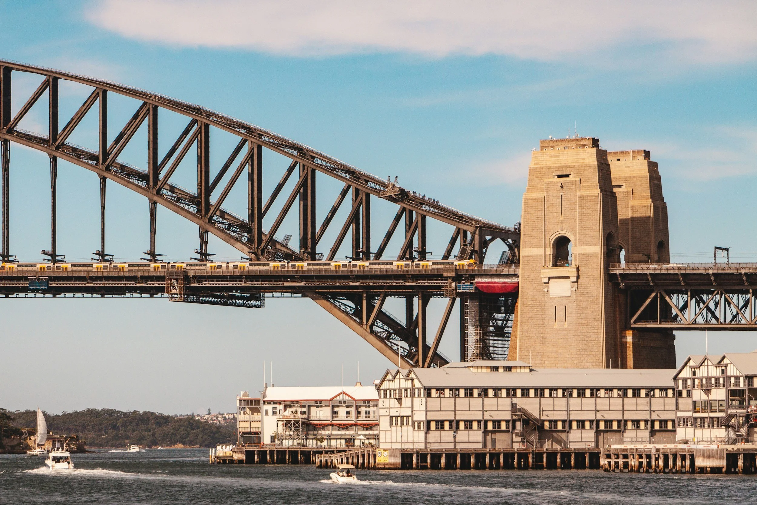 View of the Sydney Harbour Bridge with boats sailing on the water below, and buildings along the shoreline under a blue sky.